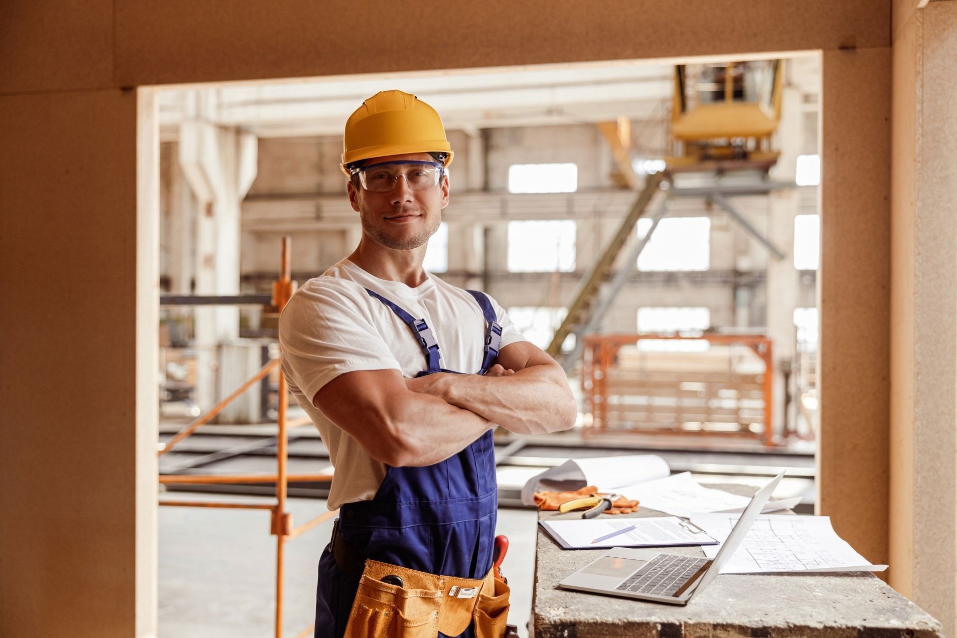 A construction worker in a yellow hard hat and blue overalls with crossed arms stands by a laptop at a building site.