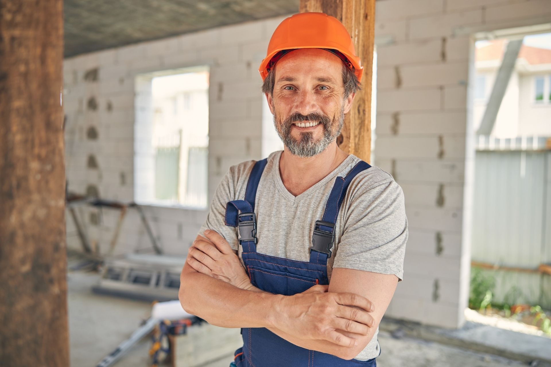 Construction worker in orange hard hat and blue overalls standing in a renovation site, smiling with arms crossed