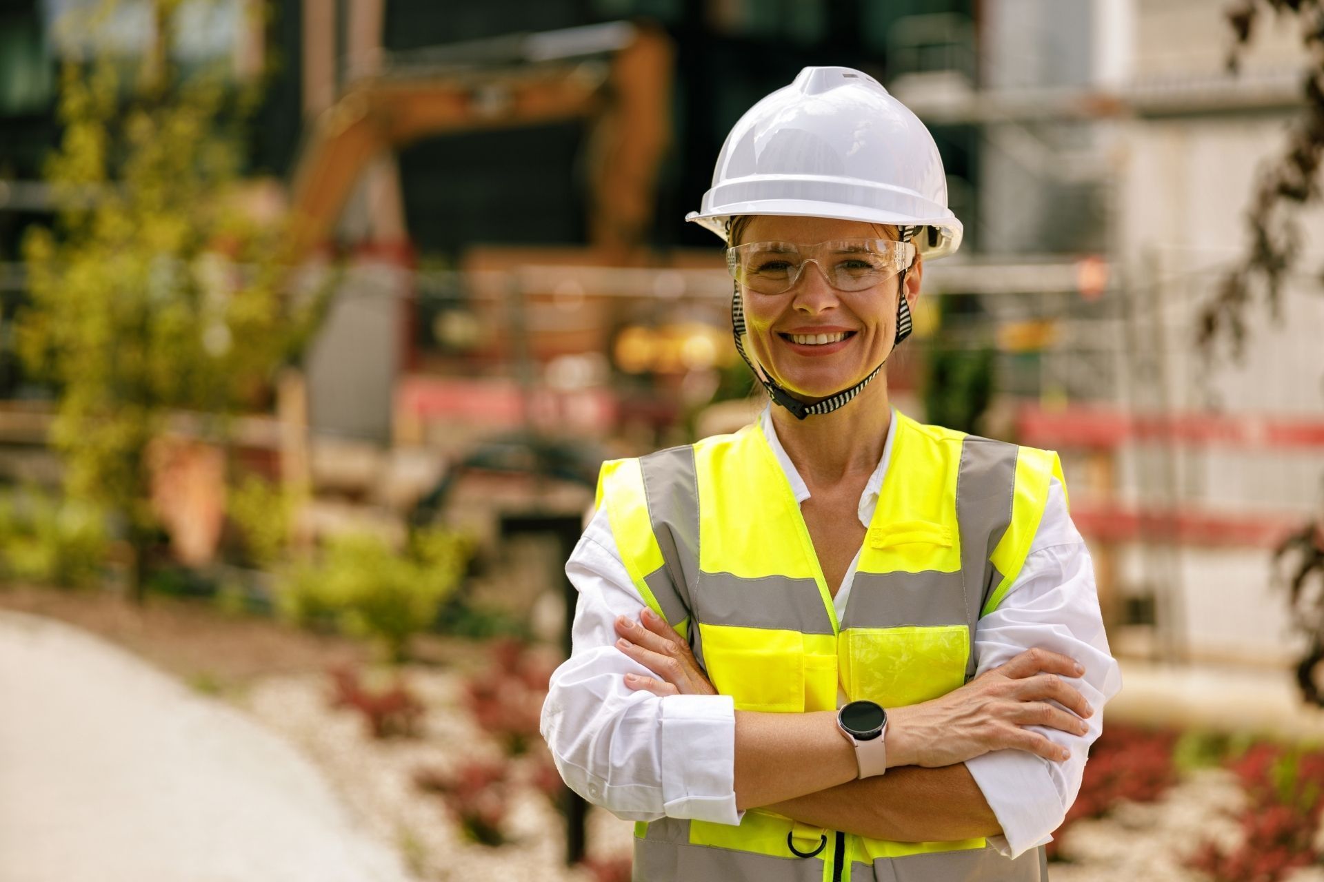 Construction worker in a white hard hat and neon vest, smiling with arms crossed at a building site