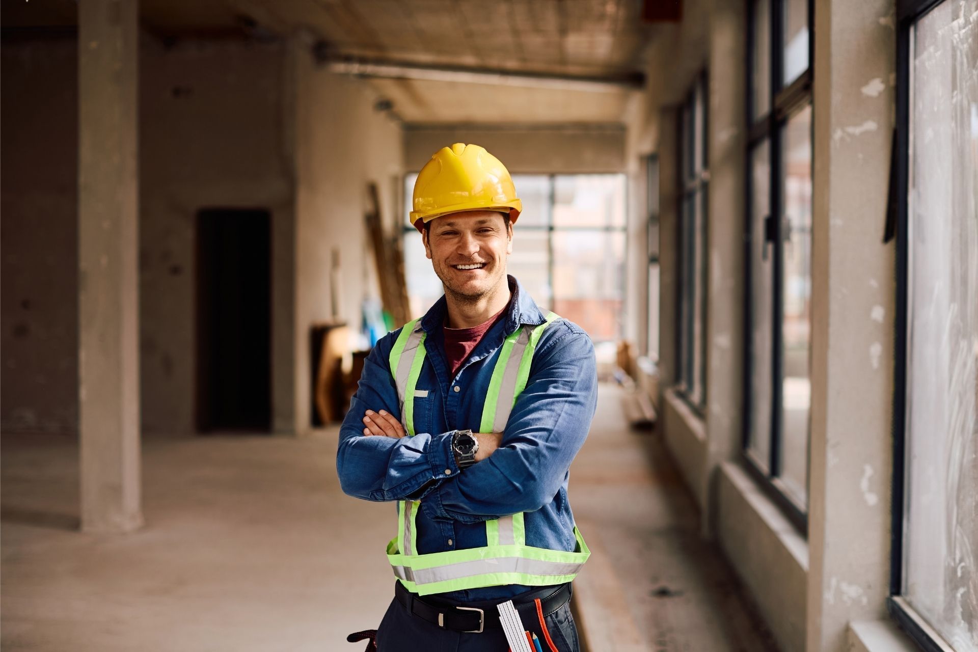 Construction worker in yellow hard hat and safety vest standing in a building under construction, smiling with arms crossed
