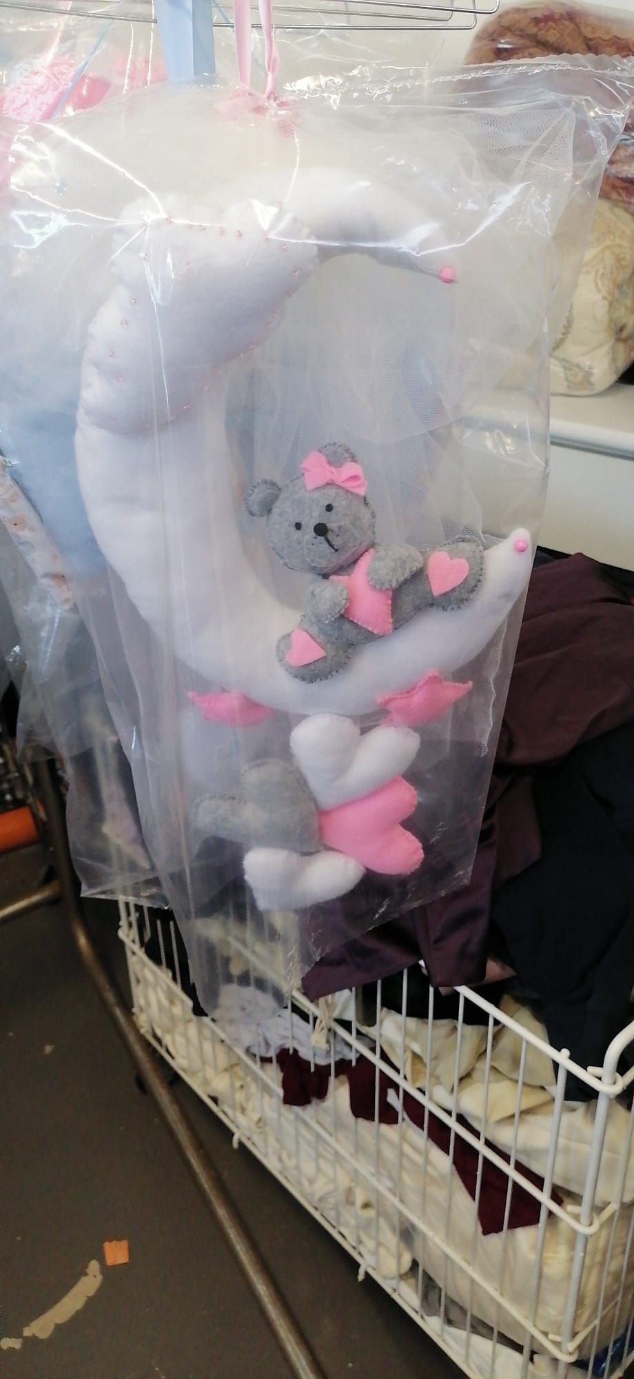 Un orsacchiotto di peluche grigio con un fiocco rosa, seduto su una mezzaluna bianca, confezionato in plastica trasparente.