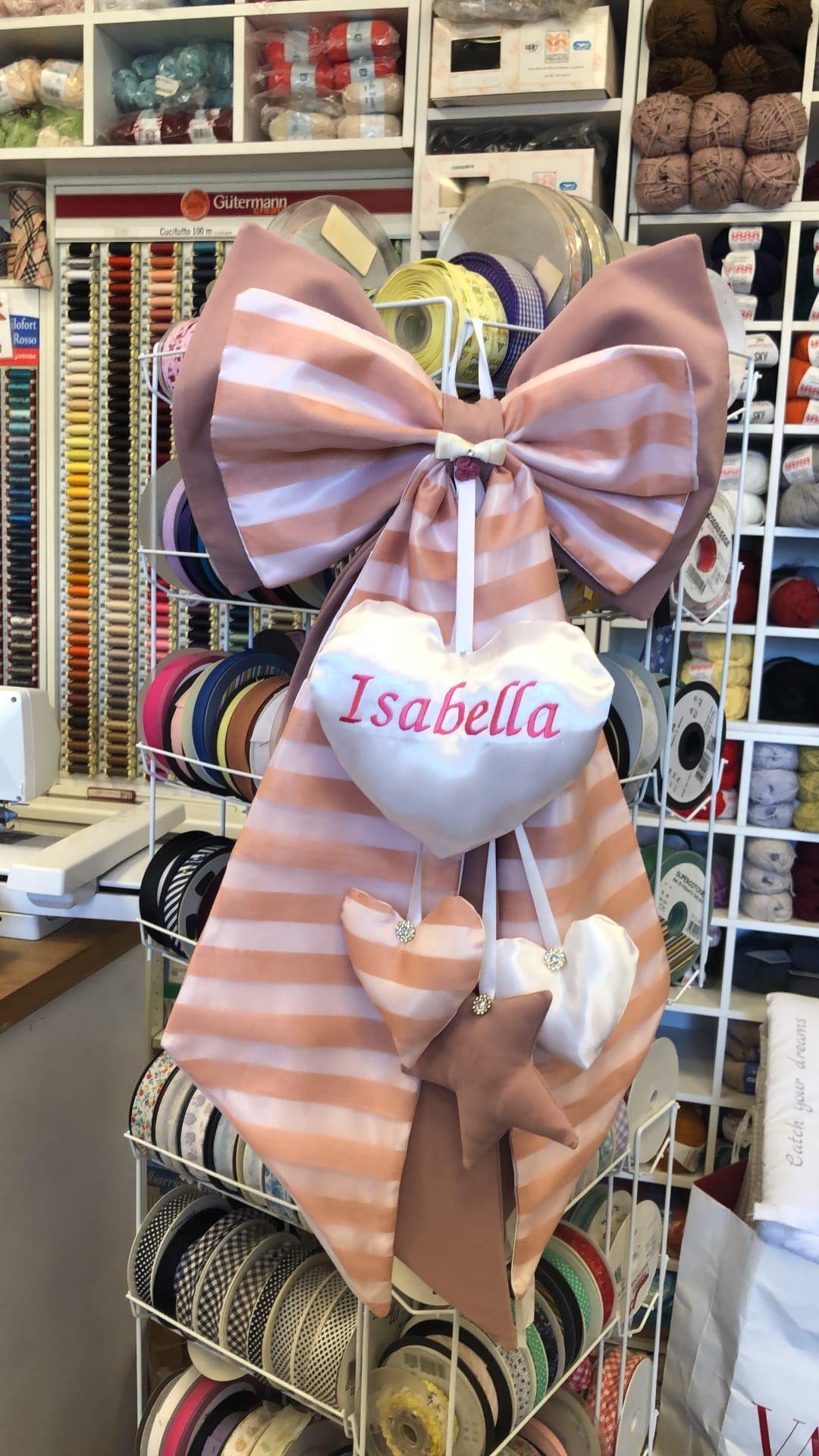 Un arazzo decorativo da parete con un grande fiocco a righe rosa e bianche, decorato con cuoricini di stoffa e una stella.