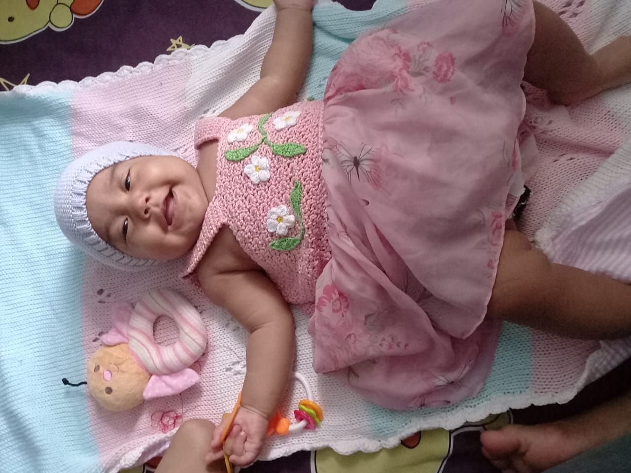 Un neonato sdraiato su una coperta colorata, con indosso un cappellino bianco e un vestitino rosa a fiori