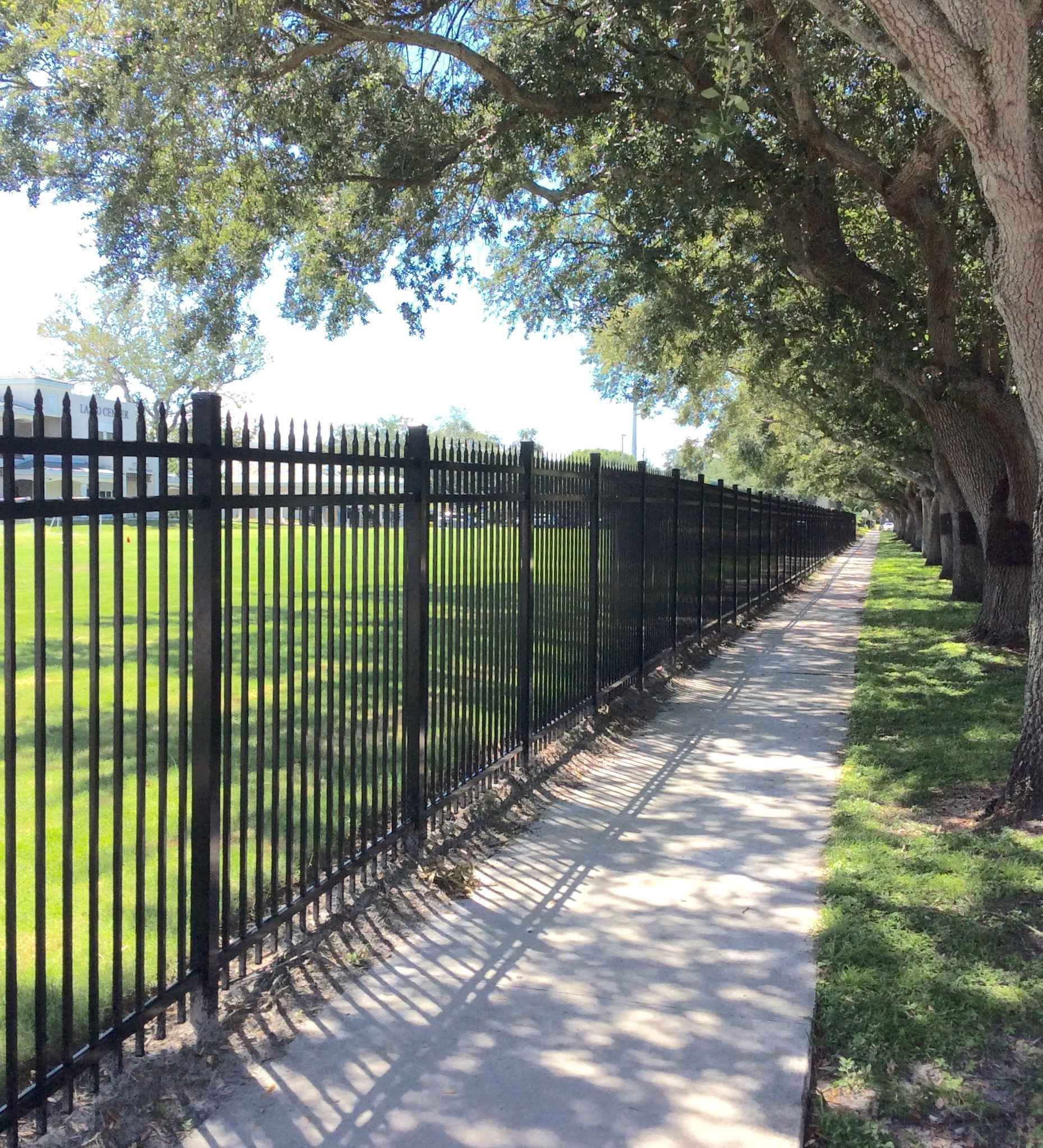 Black metal fence lines a sidewalk next to a grassy area and trees.
