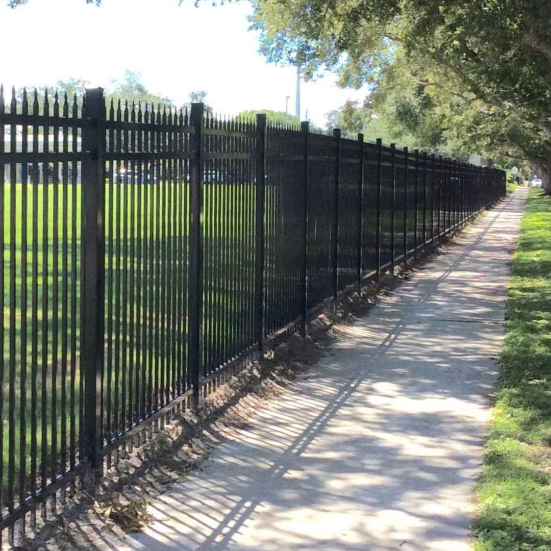 Black metal fence lines a sidewalk next to a grassy area, trees overhead.