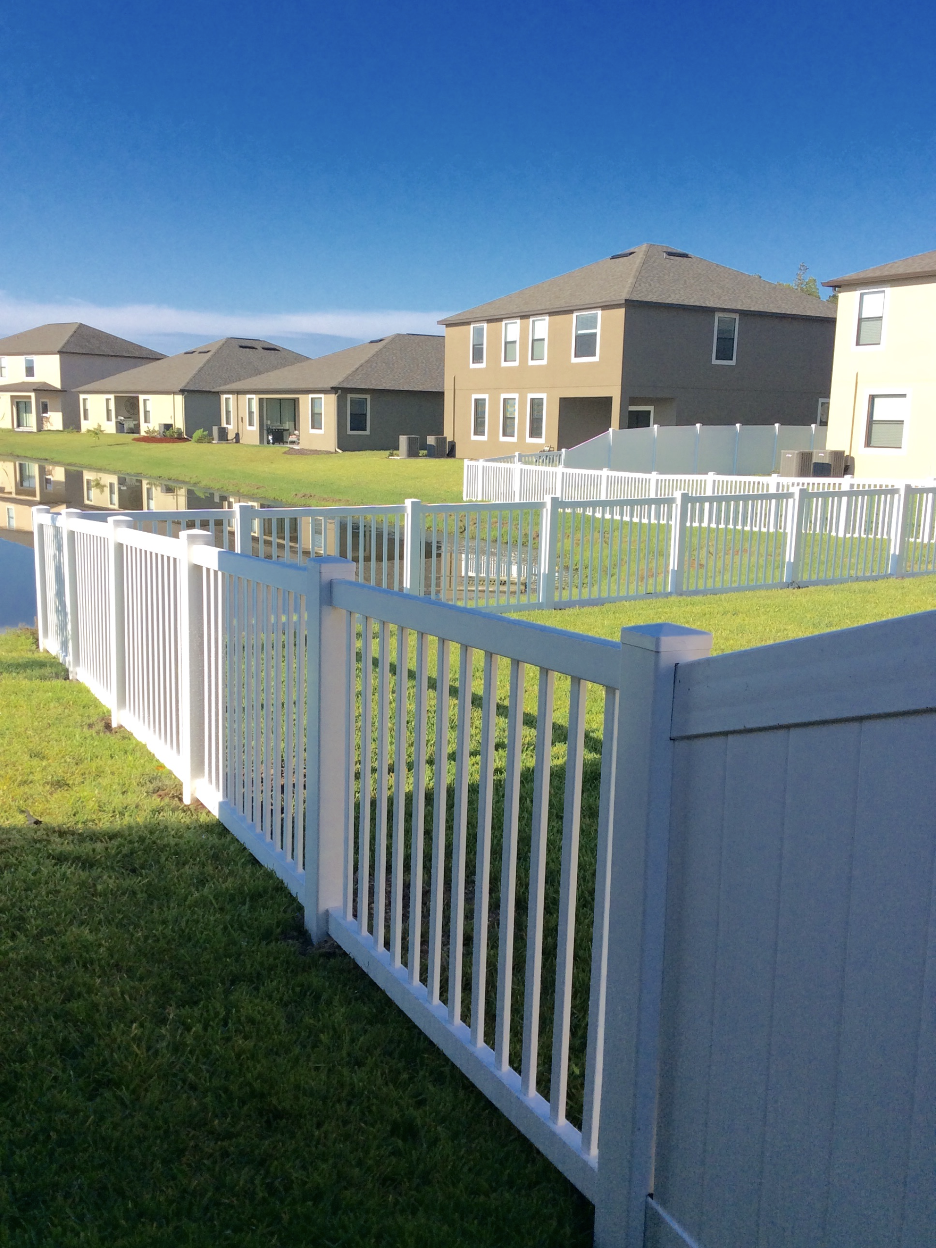 White picket fence in front yard with houses in the background. Blue sky and green grass.