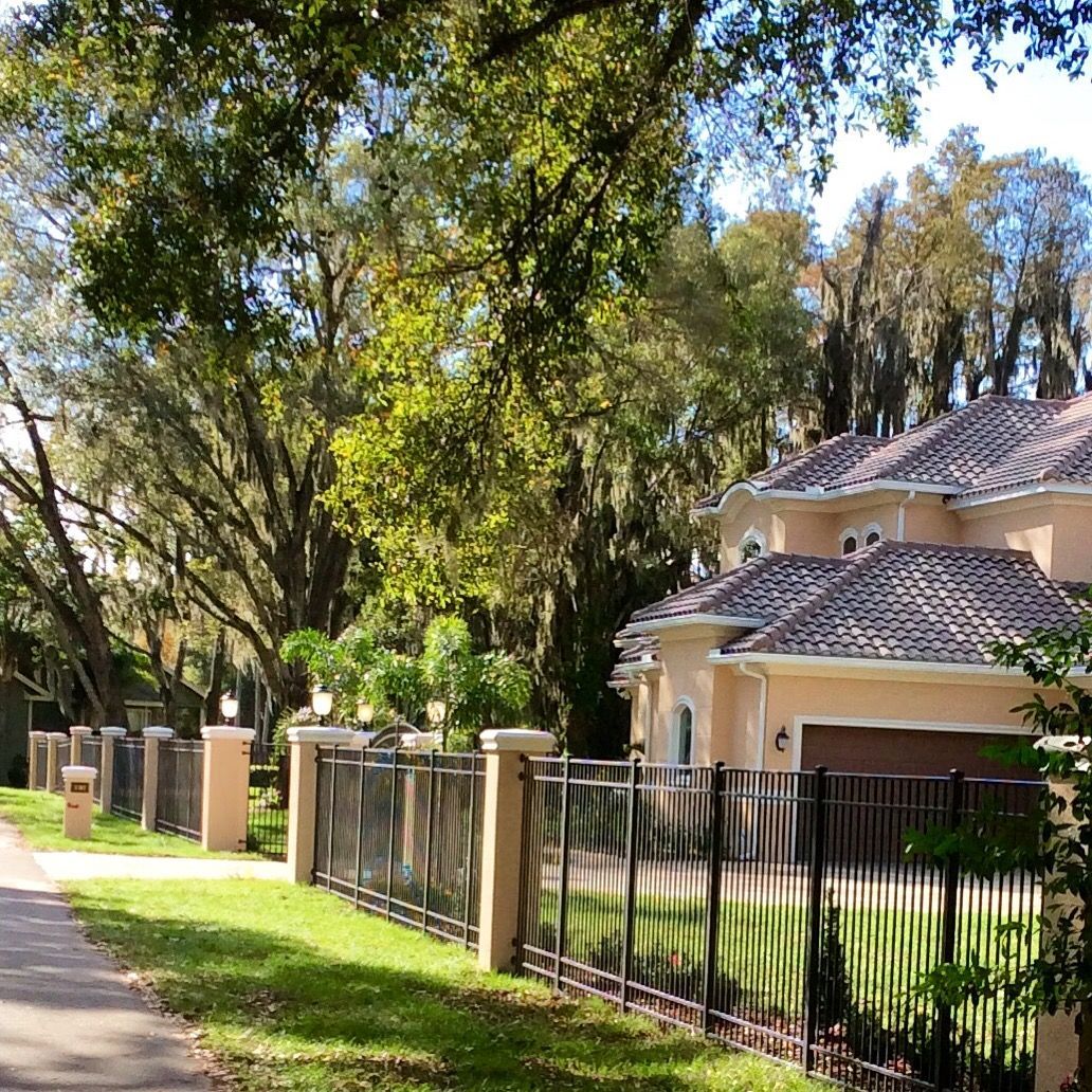 Lush yard, iron fence, and light-colored house with dark roof beneath trees, sunny day.