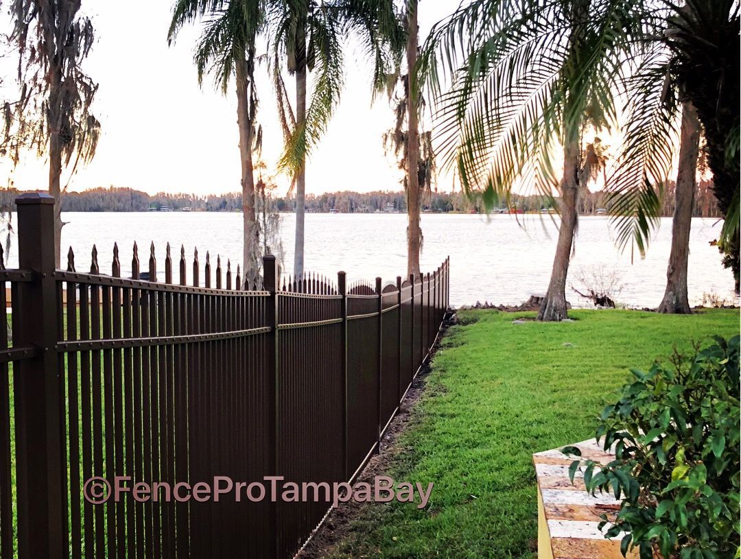 Brown metal fence along a green lawn with a lake and palm trees in the background at sunset.