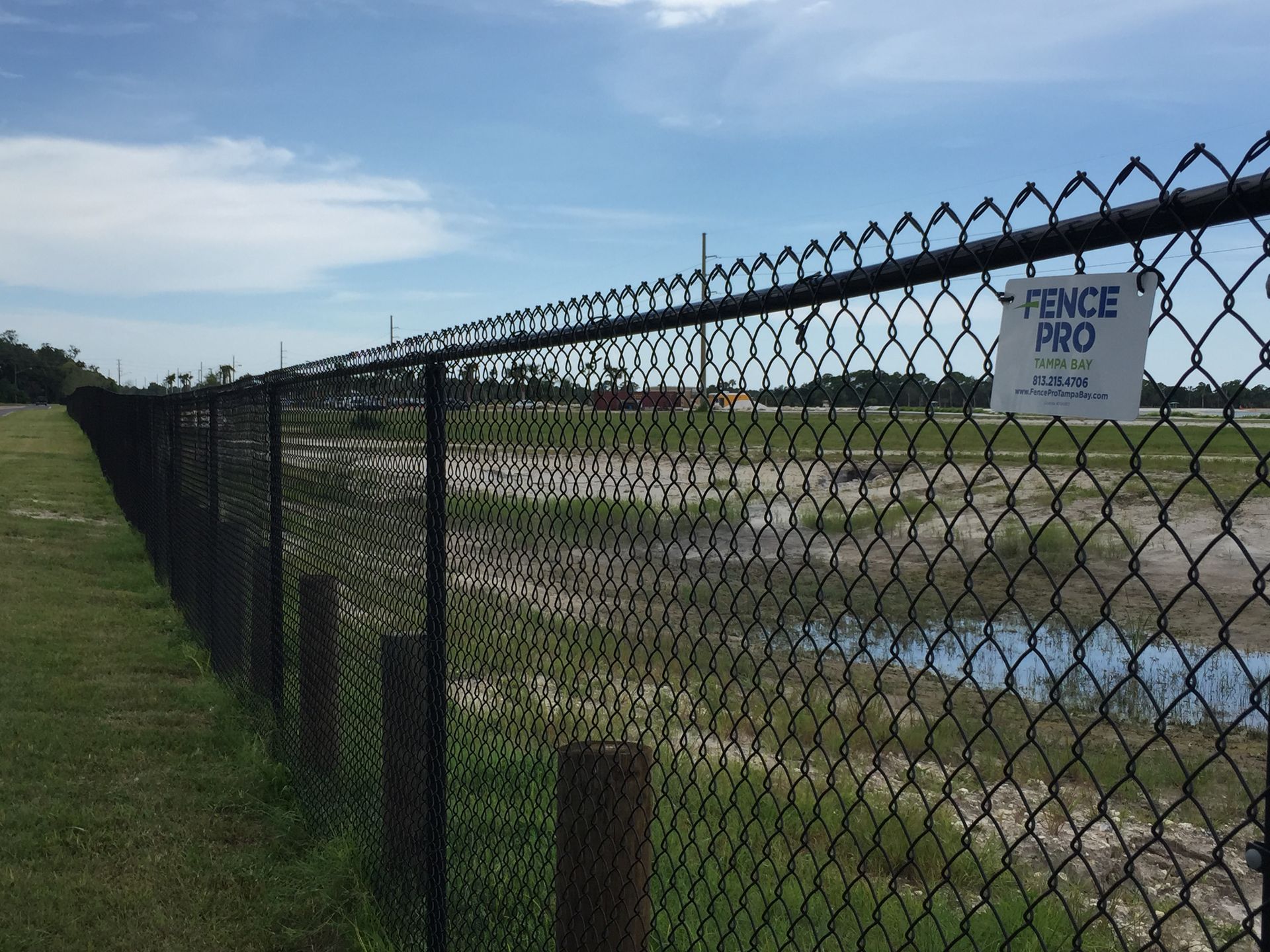 Black chain-link fence on a grassy field, with a company sign and sky in the background.
