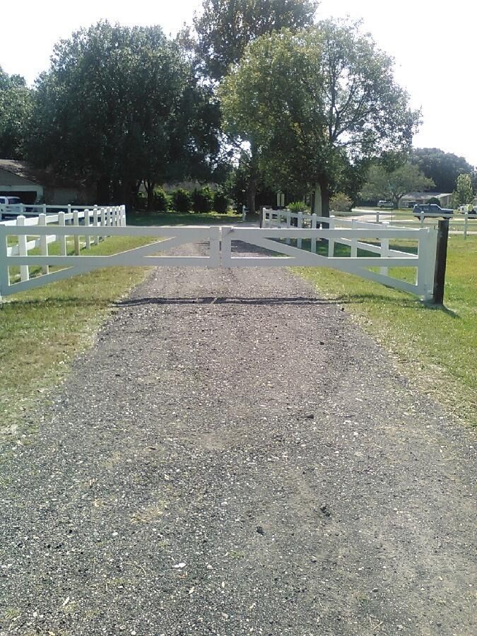 White, gated driveway entrance with gravel path, leading to a grassy area and trees under a sunny sky.