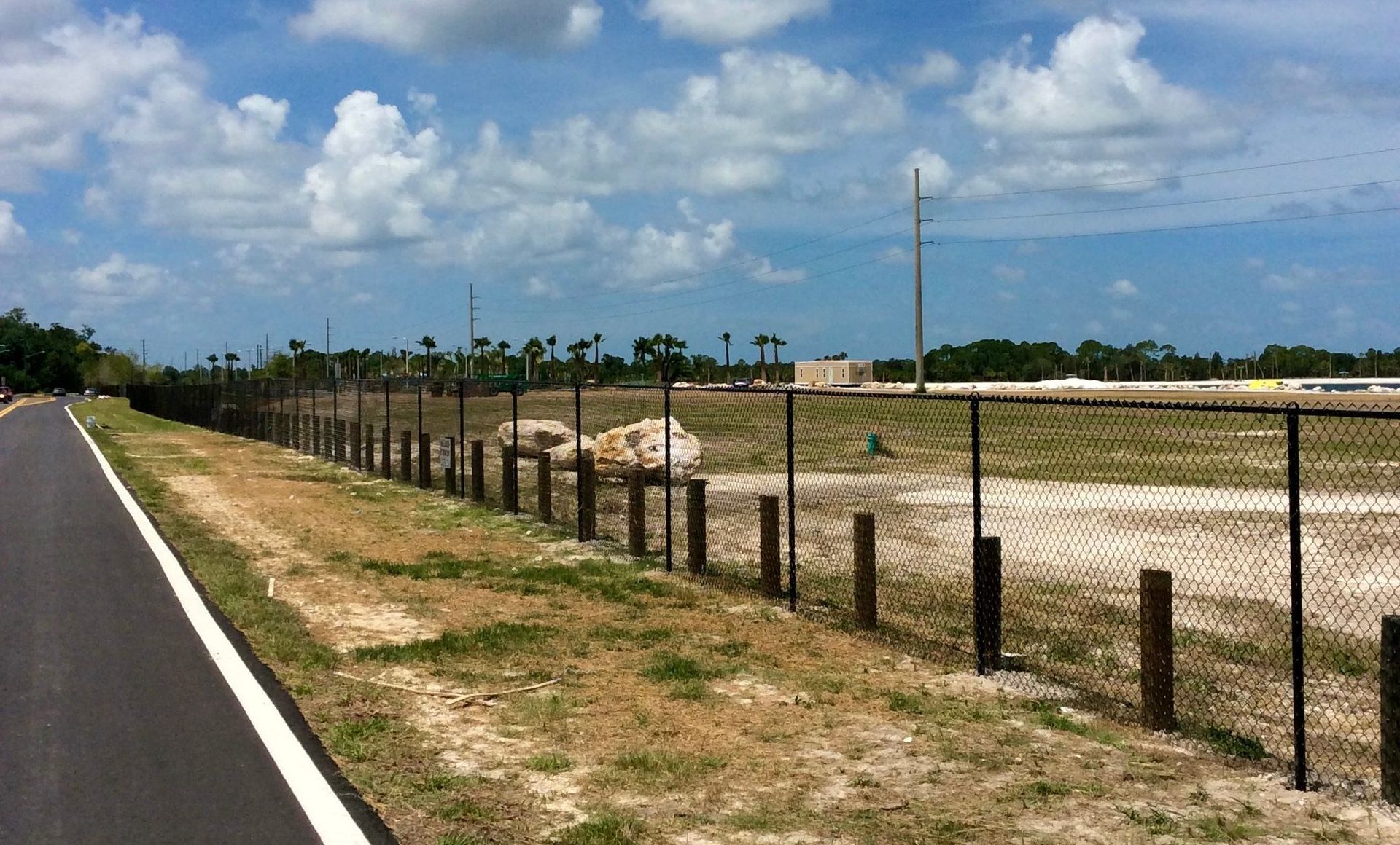 Chain-link fence borders a paved path and a vacant lot on a sunny day.