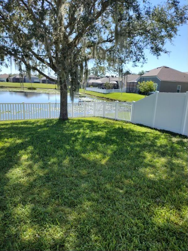 Lush green lawn with a tree casting shadows, white fence bordering a lake with homes in the background on a sunny day.