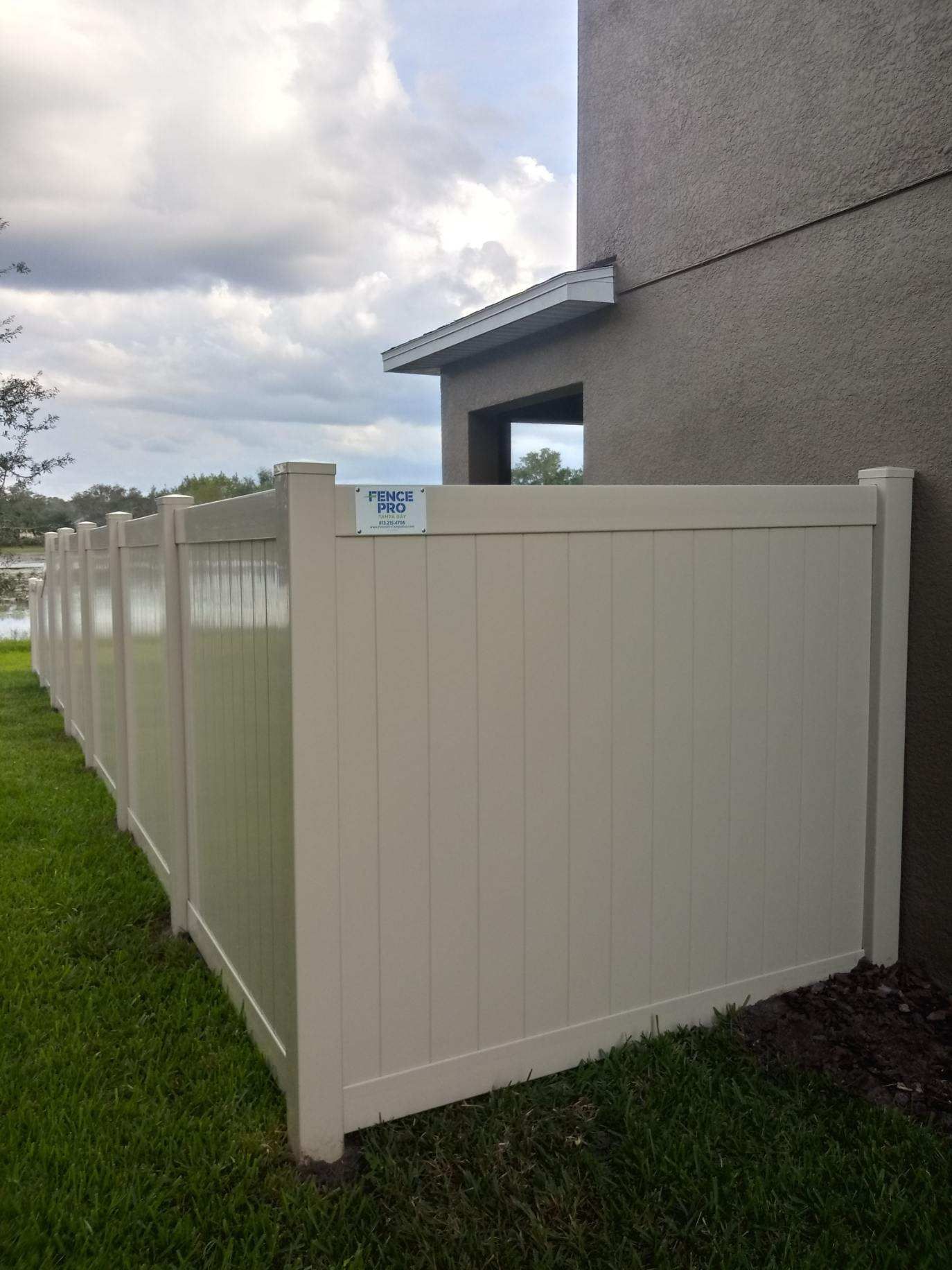 Beige vinyl fence next to a stucco building, beside grass, under a cloudy sky.