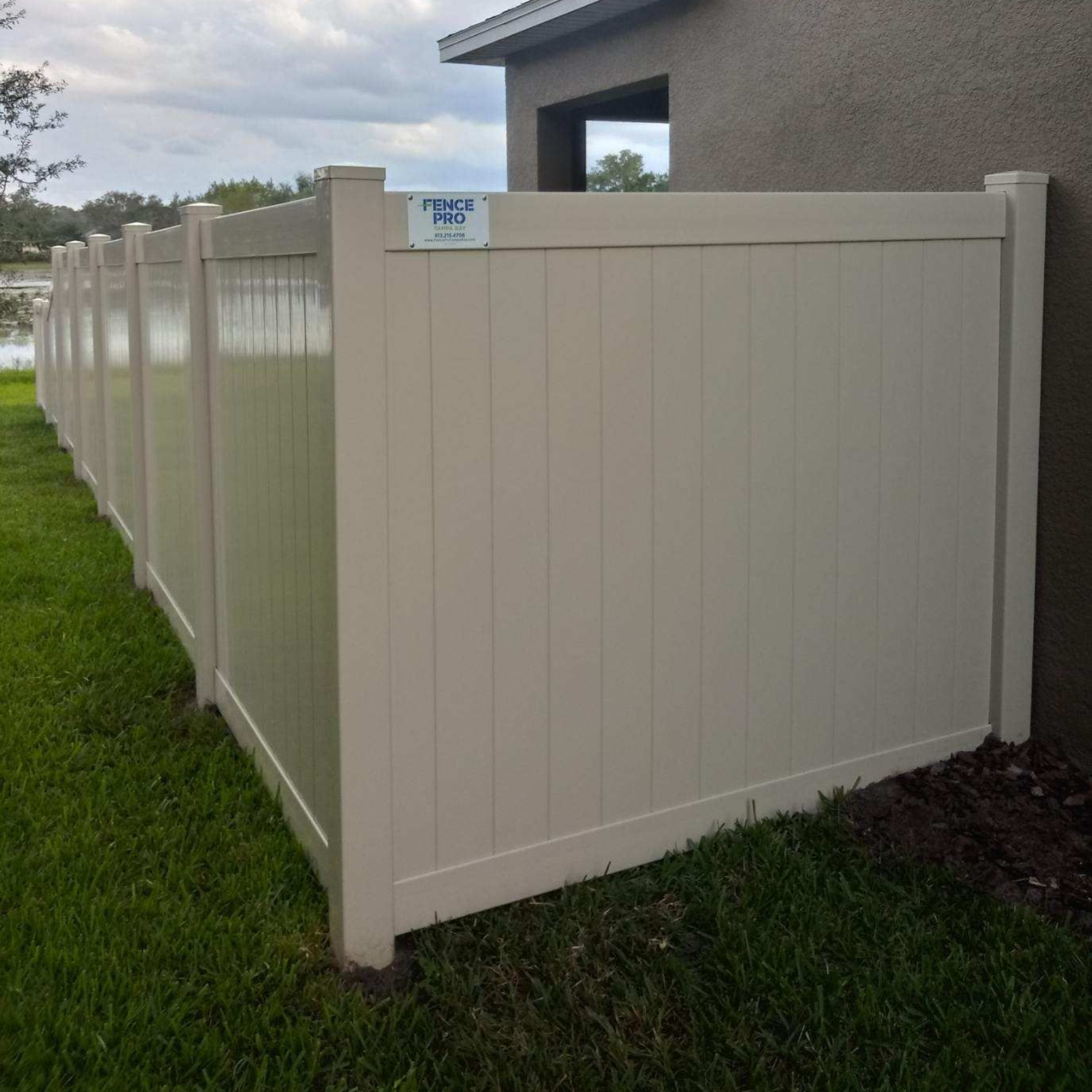 Tan vinyl fence in a grassy yard, beside a house with a window, and near water.
