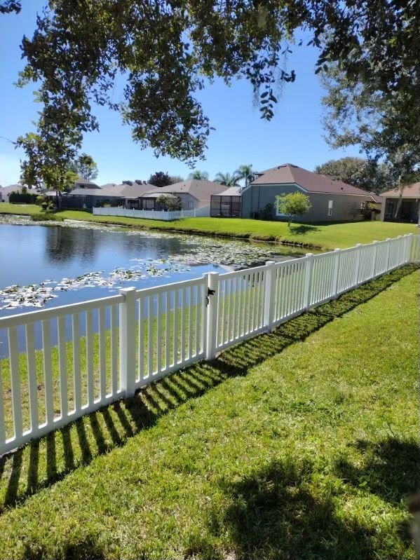 White picket fence borders a grassy yard and a pond, sunny day.