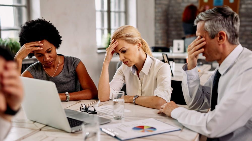 Tres compañeros de trabajo en una reunión, inclinados sobre un portátil con gráficos, con aspecto cansado y pensativo.