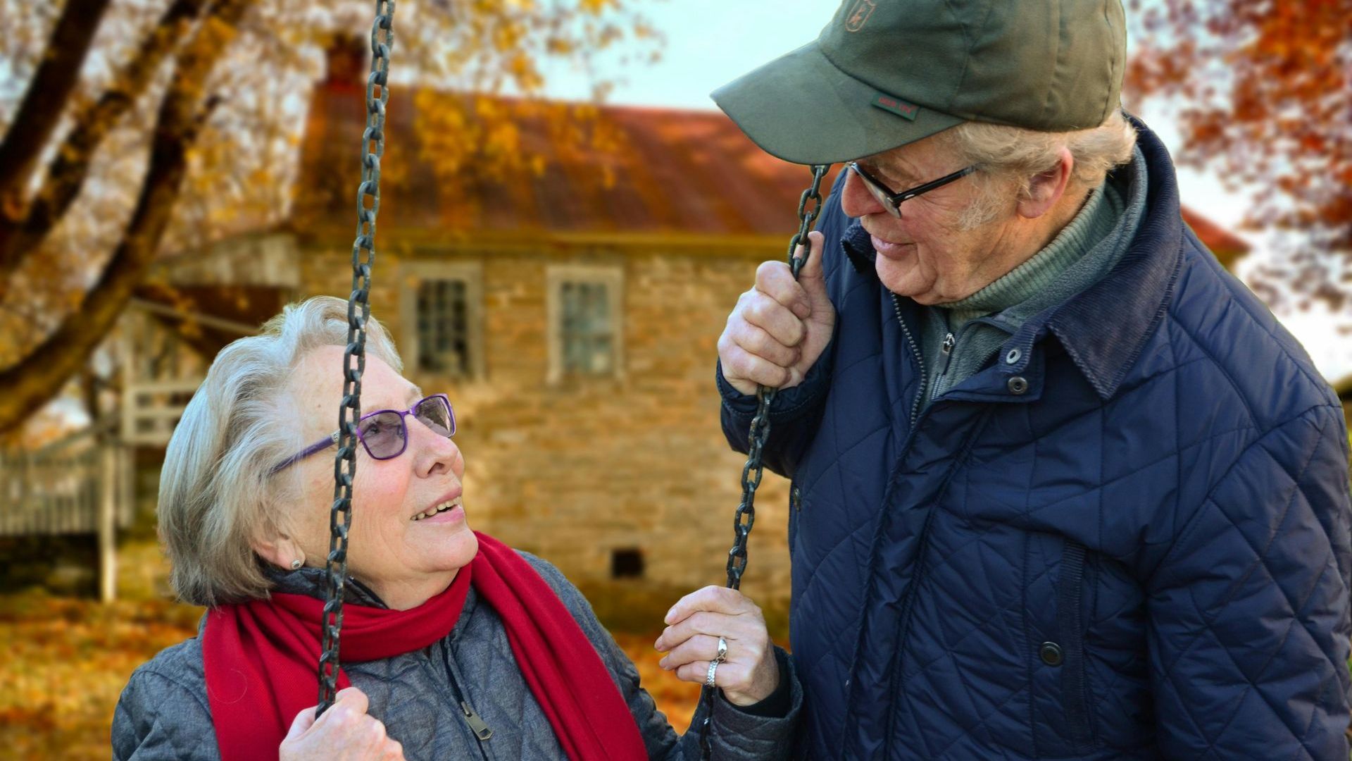 An elderly couple is sitting on a swing in front of a house.