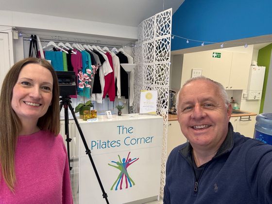 Woman and man smiling, standing near a Pilates studio sign, clothing rack in background.