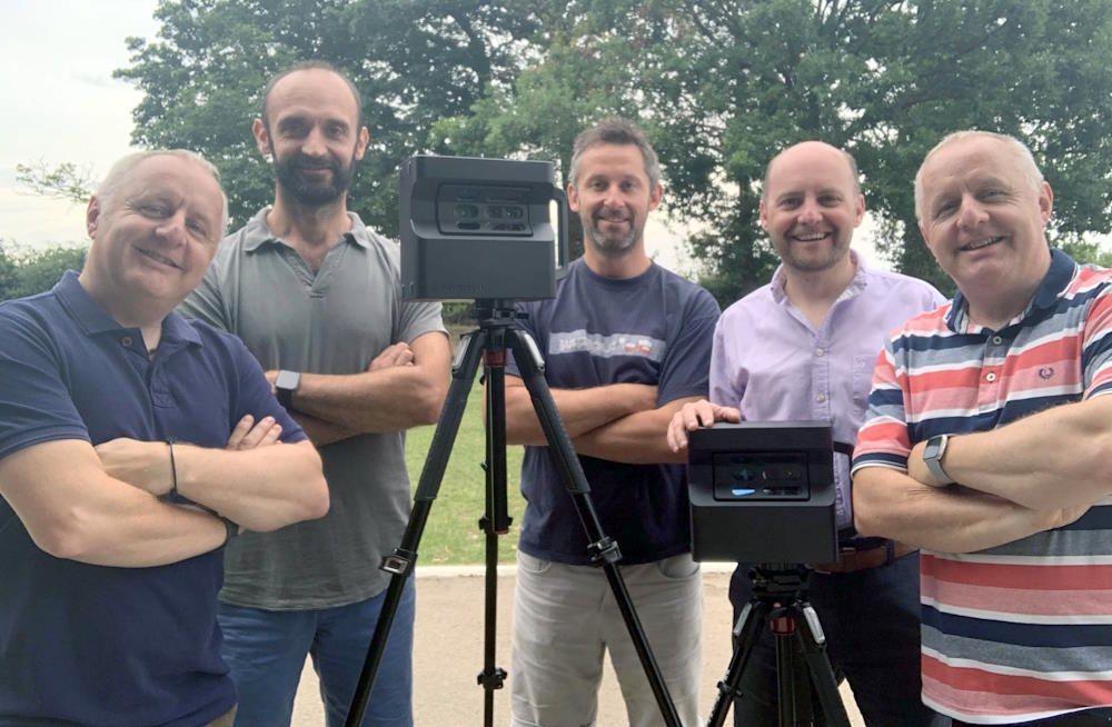 A group of men are standing in front of a camera on a tripod.