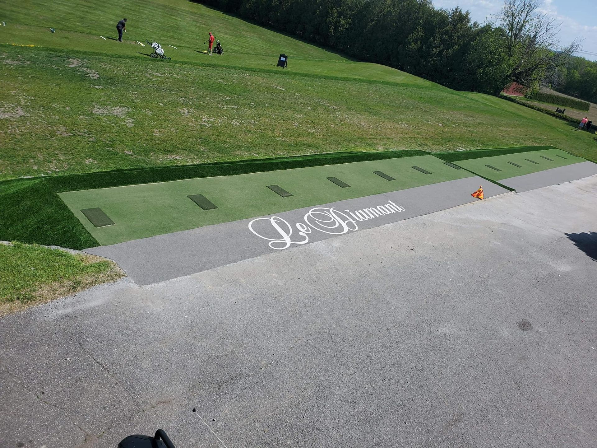 Practice de golf avec une zone de frappe verte, une allée grise et le logo « Le Dimond ». Des personnes sont visibles sur l'herbe en arrière-plan.