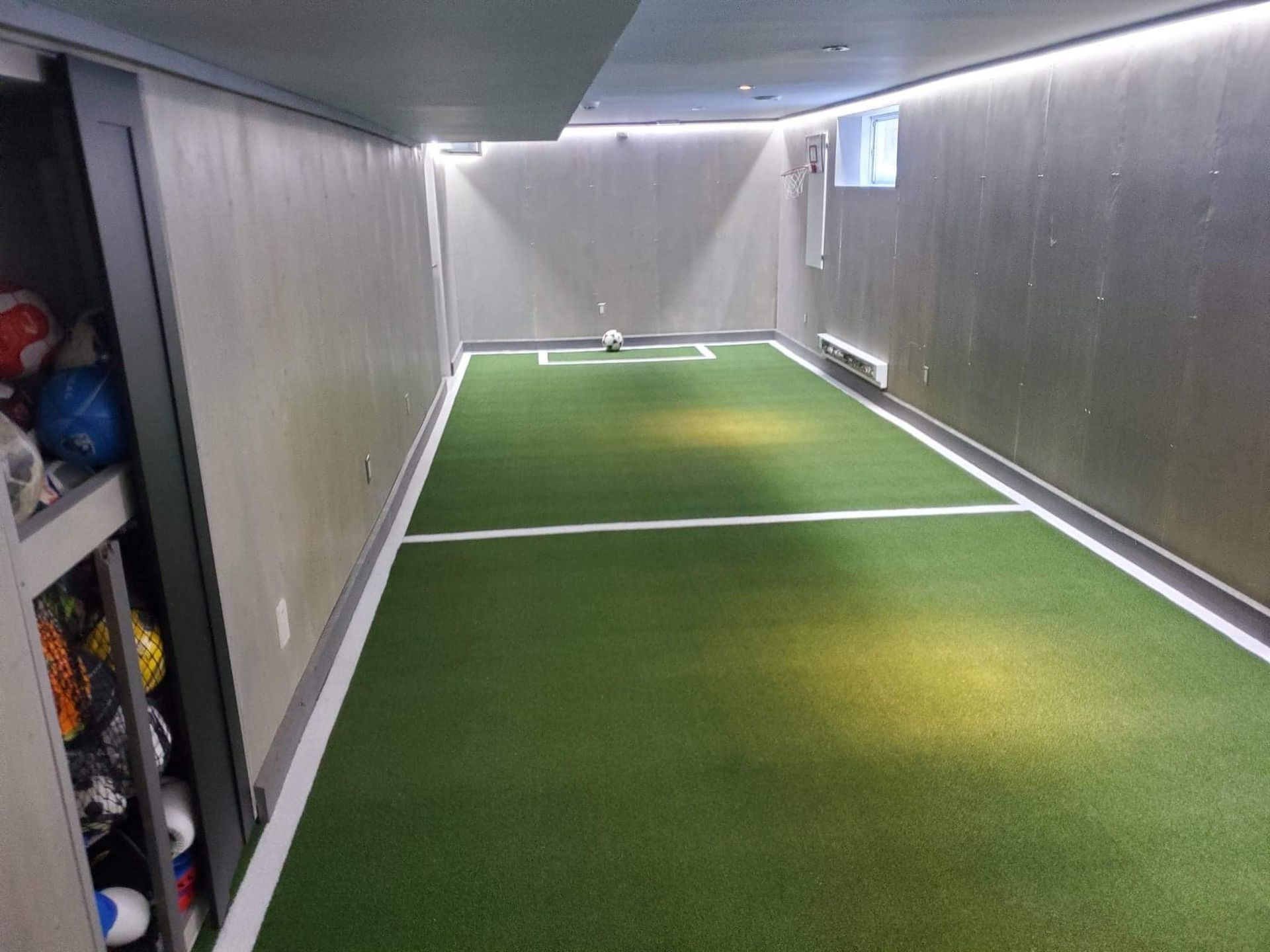 Un petit terrain de football intérieur avec gazon vert, lignes blanches et but. Un espace de rangement pour les ballons et le matériel se trouve à gauche.