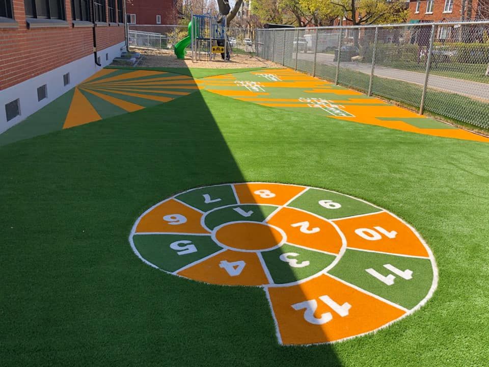 Une aire de jeux en gazon vert avec une marelle colorée et d'autres aires de jeux. Le bâtiment en briques se trouve à gauche et une clôture borde celle de droite.