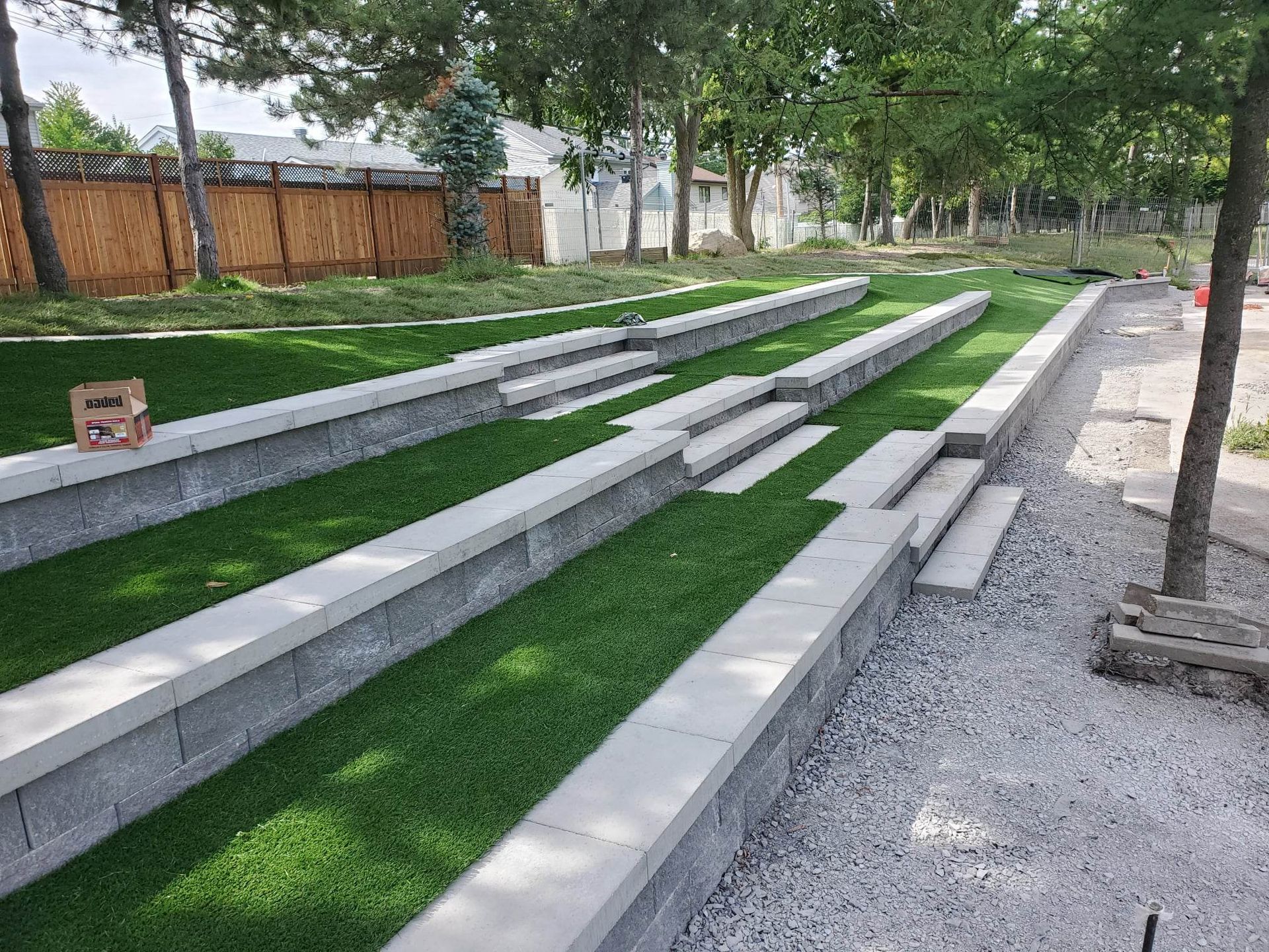 Espace extérieur avec gradins et gazon artificiel, murs en blocs gris et petits escaliers, entouré d'arbres et de gravier.