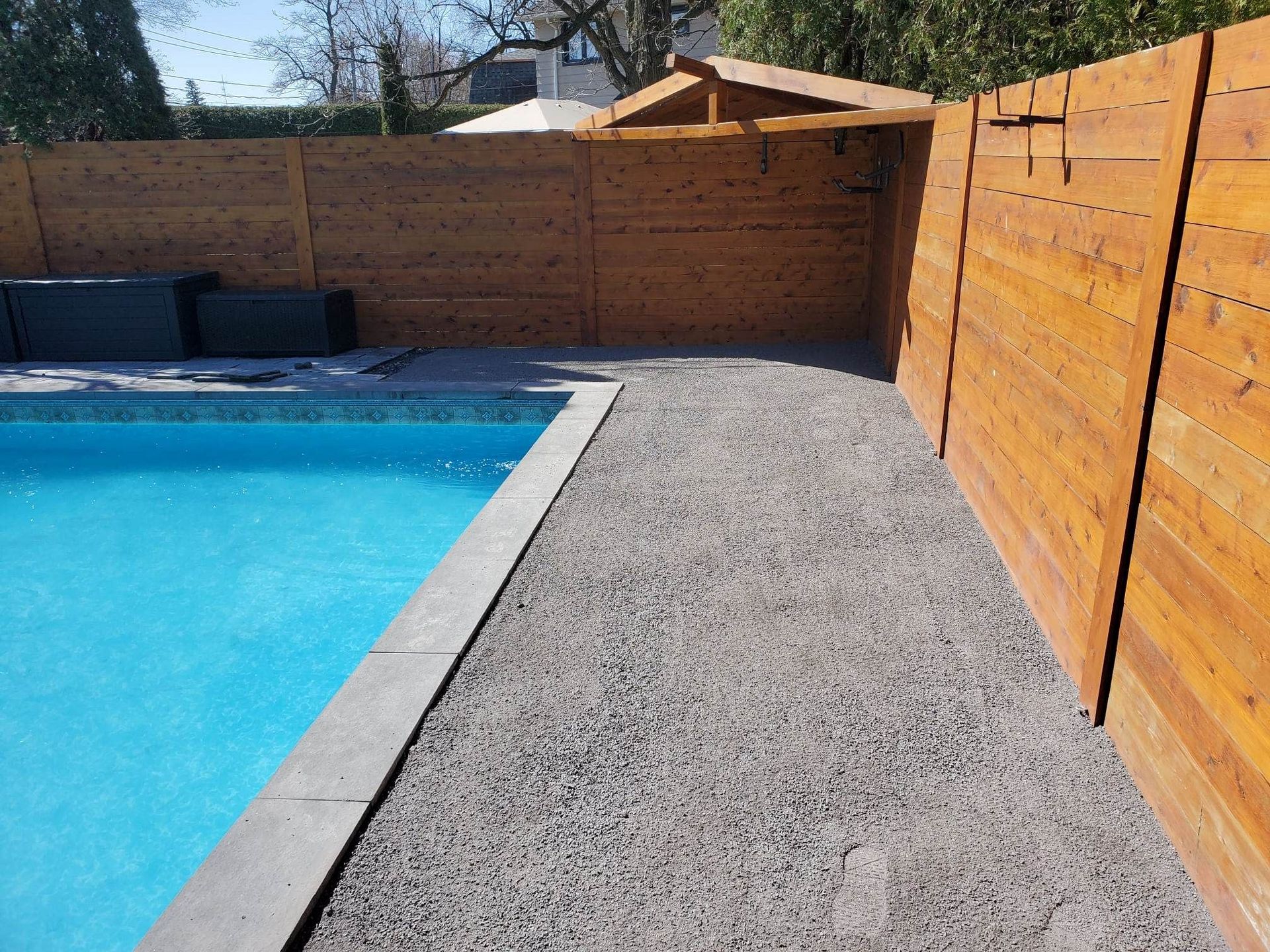 Un jardin avec une piscine bleue, un chemin de gravier et une clôture en bois teinté. Un petit abri en bois se trouve à l'arrière-plan.