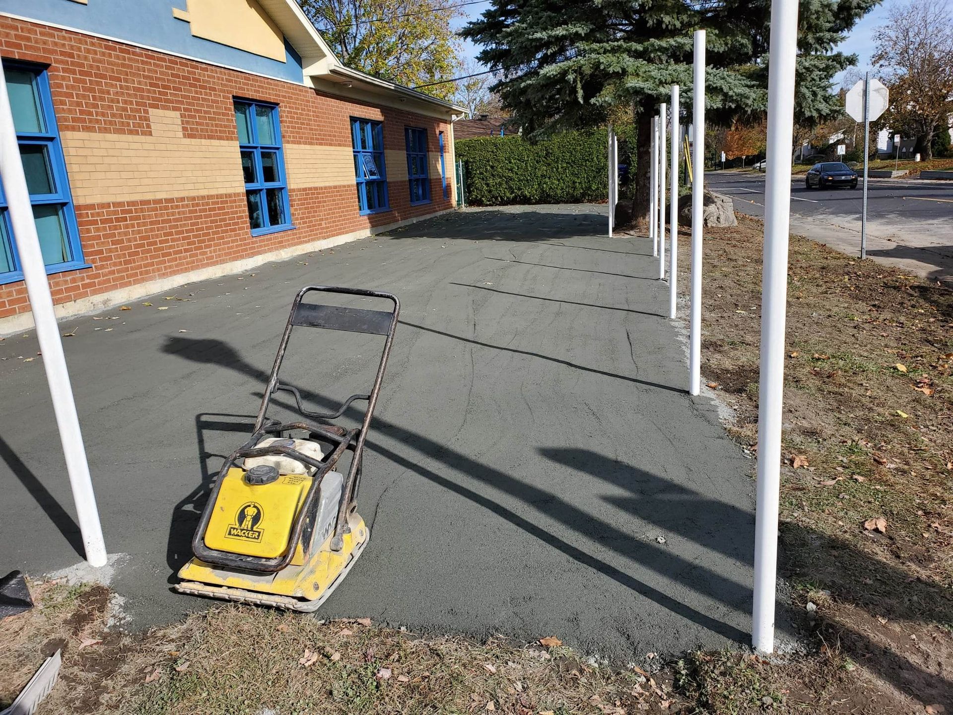 Allée asphaltée avec un compacteur jaune. Des poteaux blancs bordent le côté, à côté d'un bâtiment en briques rouges.