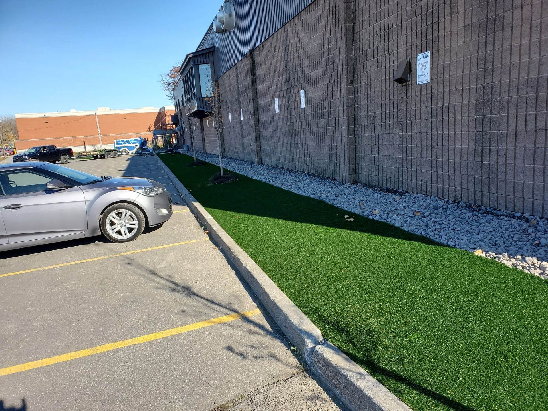 Une voiture grise garée sur un terrain à côté d'un bâtiment avec une bande de gazon artificiel et des rochers.