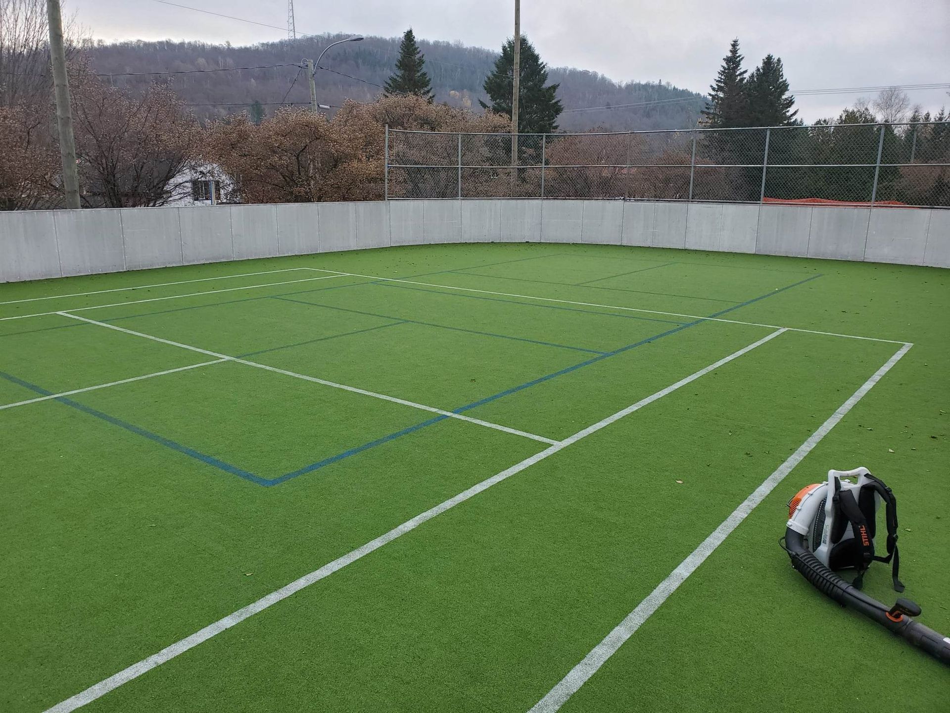 Un terrain de sport en gazon artificiel vert avec des lignes blanches et bleues, un souffleur de feuilles et un arrière-plan d'arbres et de collines.