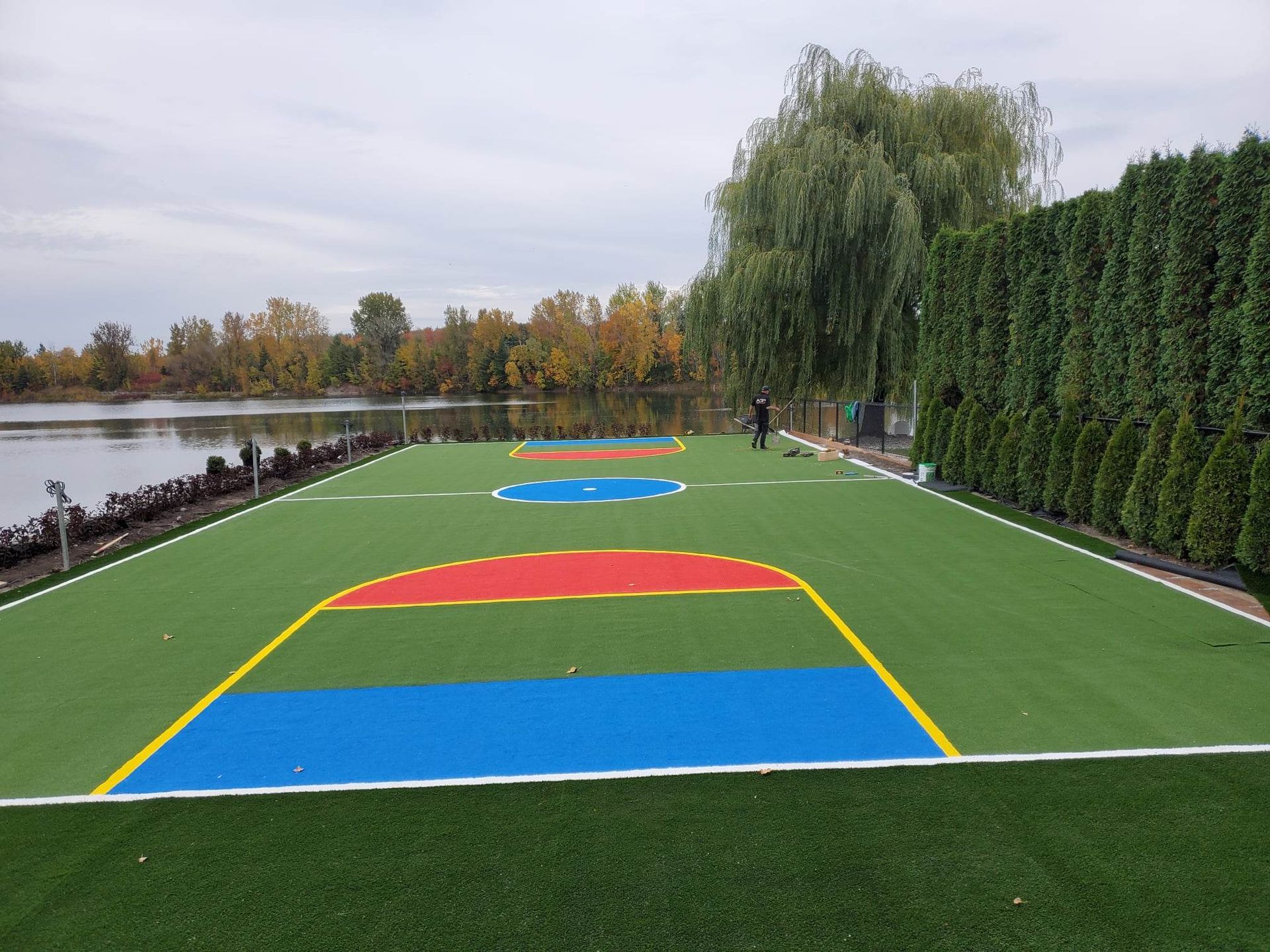 Un terrain de basket coloré sur une pelouse au bord d'un lac. Le terrain est composé de sections bleues, rouges et jaunes et borde un plan d'eau.