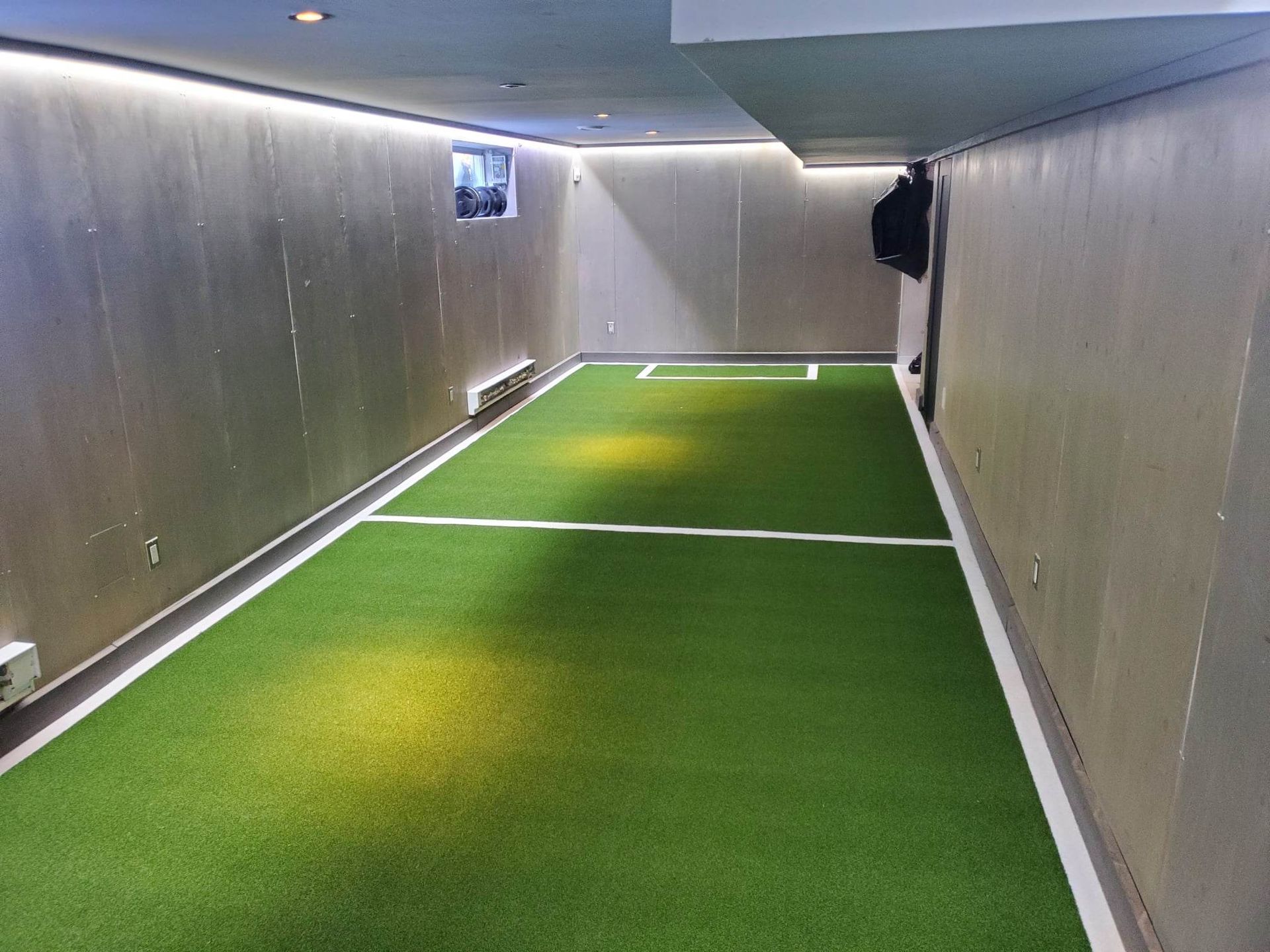 Terrain de mini-football intérieur en gazon vert et lignes blanches, éclairé par des spots. Des murs gris entourent l'aire de jeu.