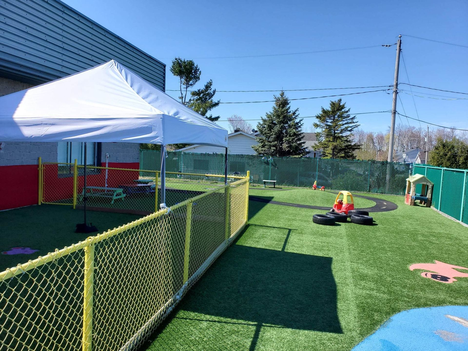 Aire de jeux avec gazon vert, clôture grillagée jaune, auvent blanc et équipements de jeu sous un ciel ensoleillé.