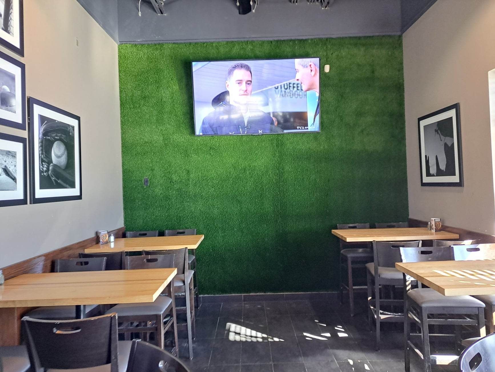 Un intérieur de restaurant avec des tables, des chaises et un téléviseur fixés sur un mur en gazon artificiel vert. Des photos encadrées en noir et blanc sont exposées sur le mur de gauche.