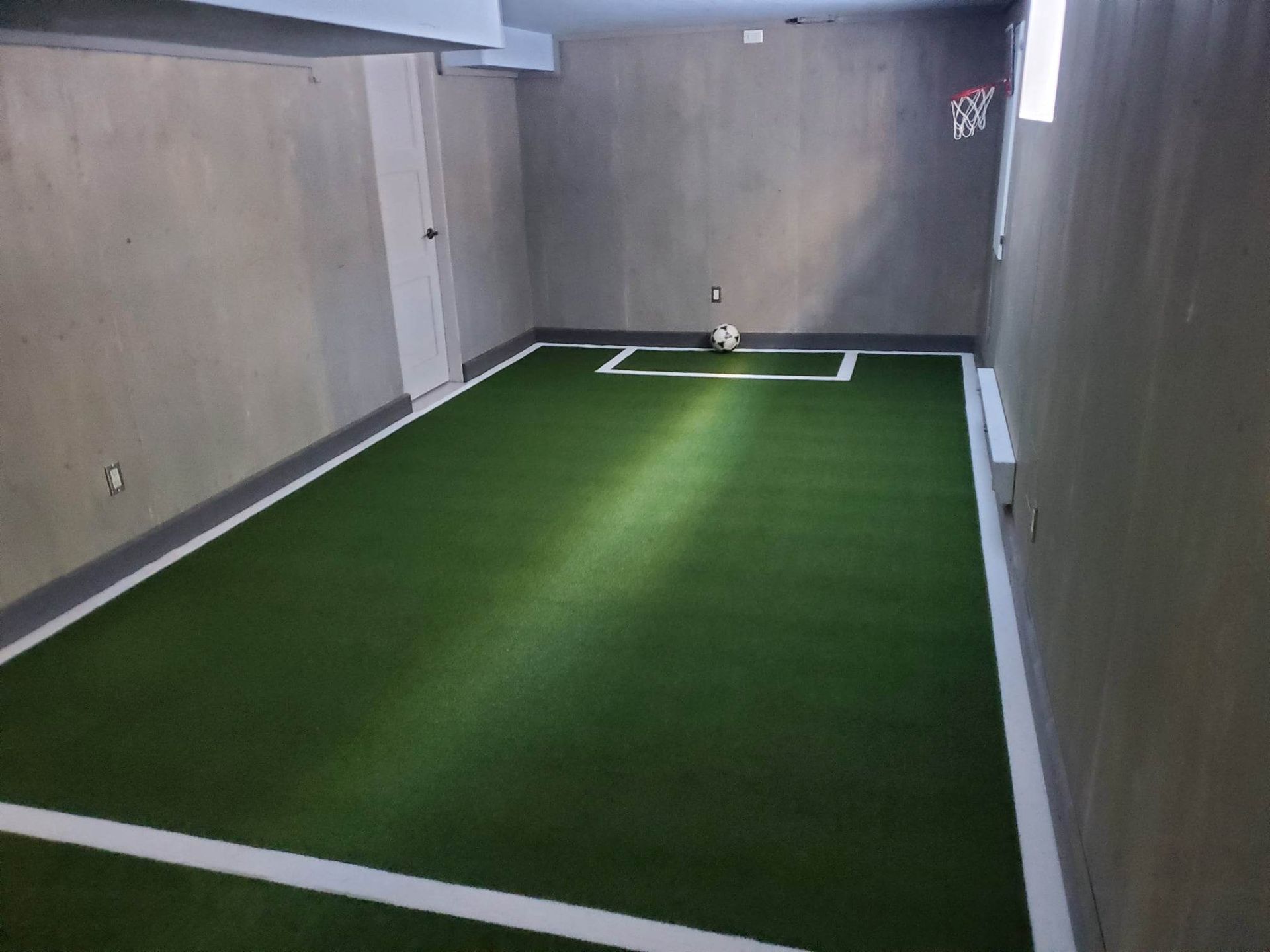Terrain de football intérieur en gazon synthétique, lignes blanches et ballon. Des murs gris entourent l'aire de jeu.
