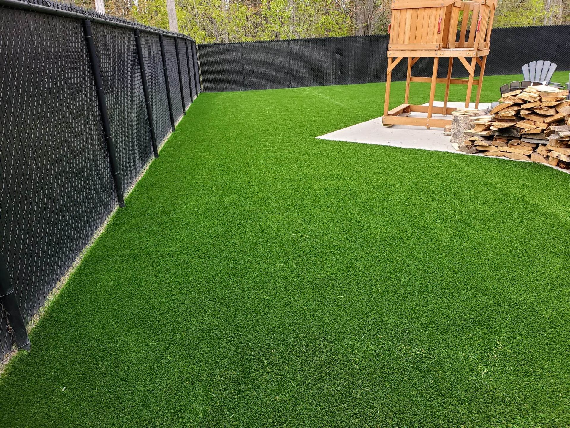 Une cour clôturée avec du gazon artificiel vert vif, une structure de jeu en bois et un tas de bois.