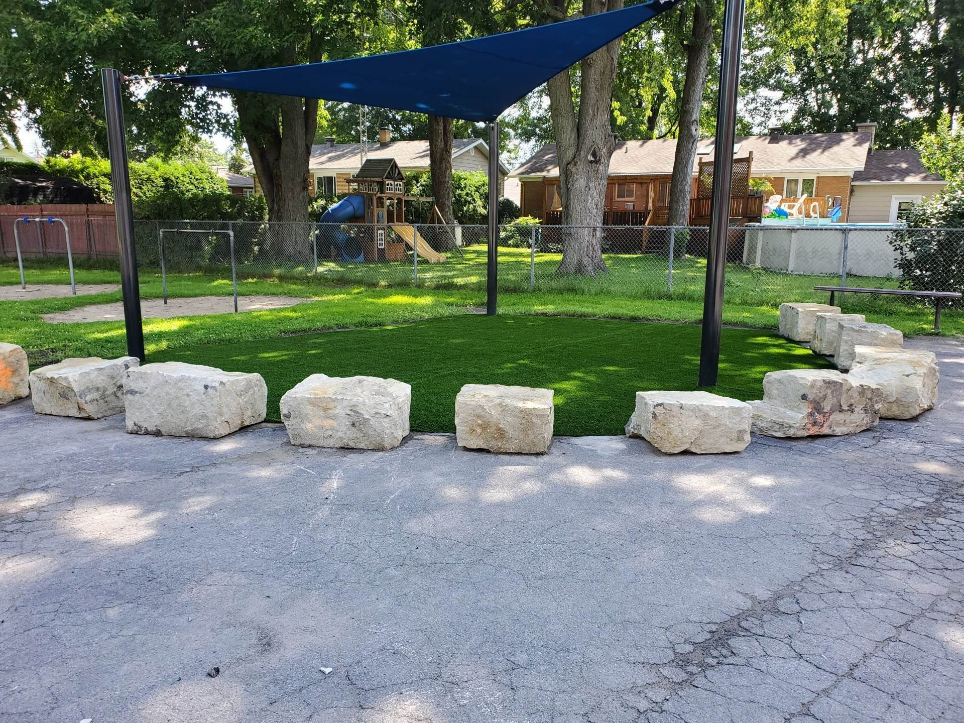 Un espace extérieur ombragé avec un cercle de blocs de pierre sur l'herbe. Un auvent bleu et des arbres dominent le paysage, et une aire de jeux se dessine en arrière-plan.