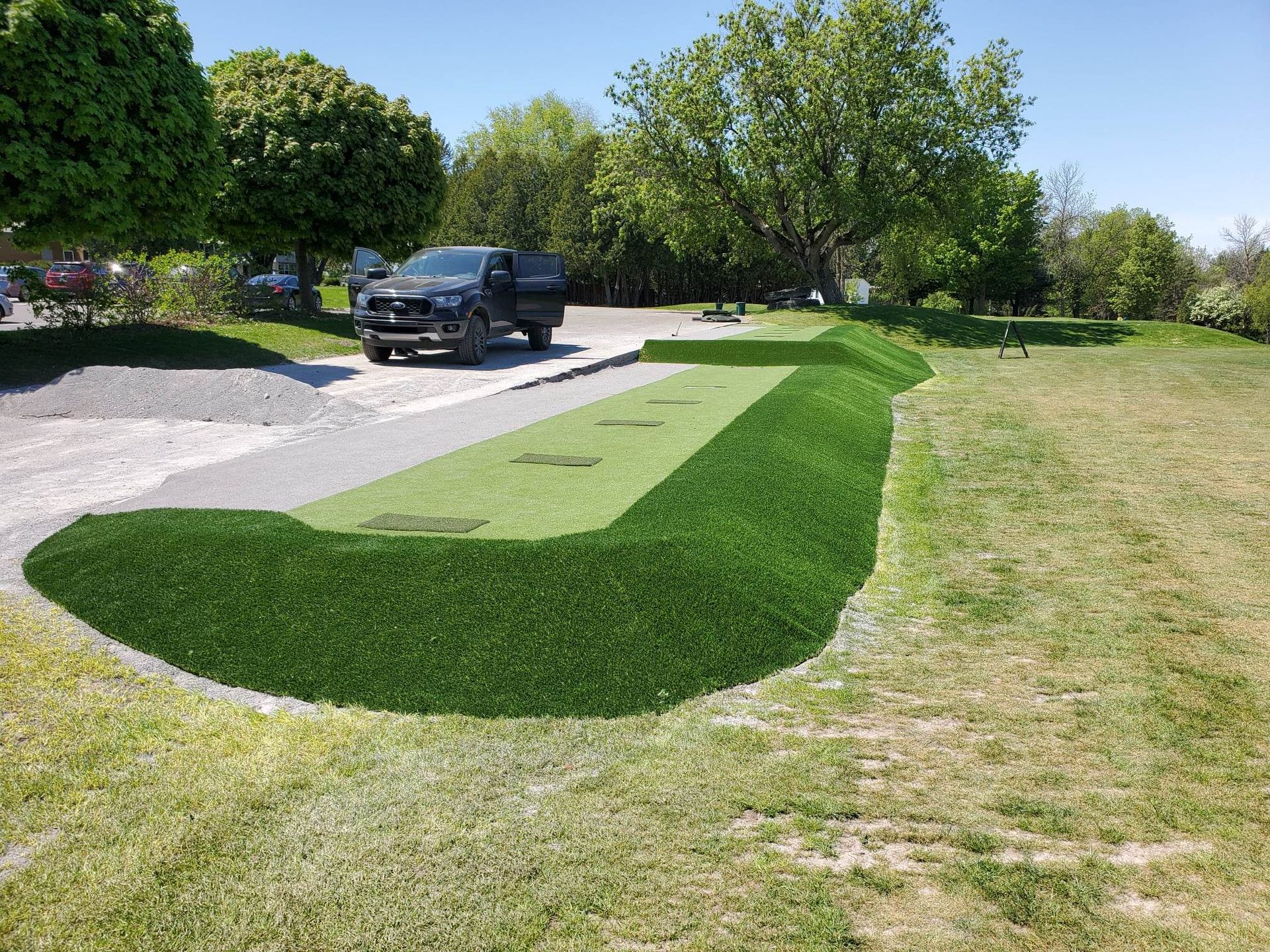 Un camion noir garé sur du gravier à côté d'une bande de gazon artificiel et d'herbe, avec un ciel clair au-dessus.
