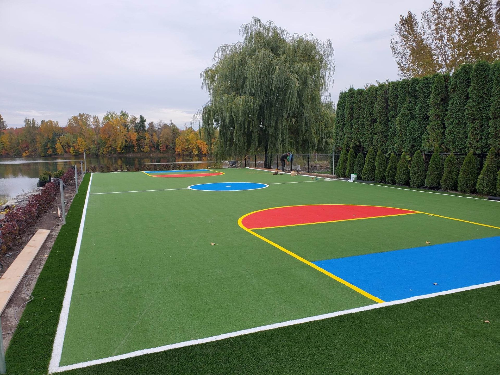 Terrain de sport extérieur avec une surface en gazon artificiel vert, comportant des marquages colorés pour divers jeux, à côté d'un lac avec des arbres.
