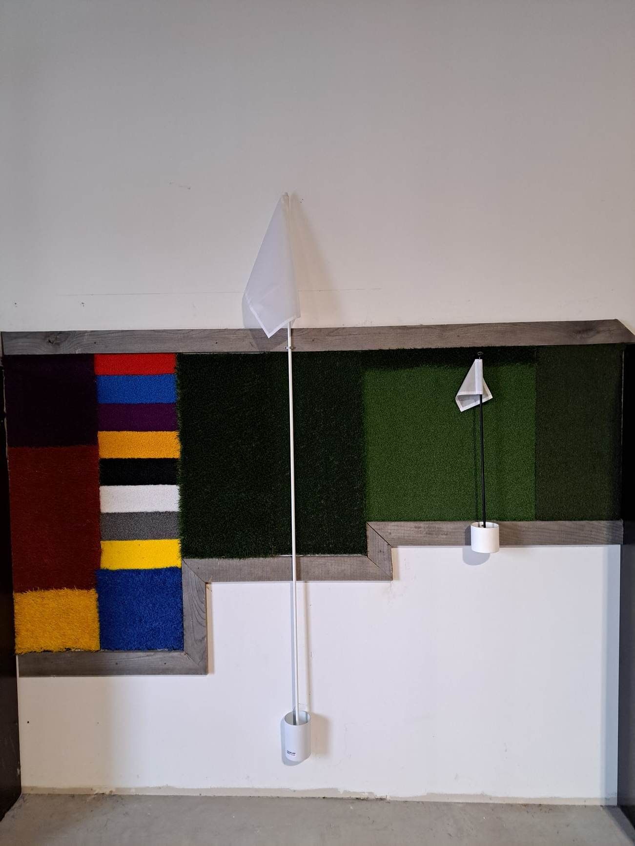 Installation de mini-golf intérieur avec un drapeau blanc et du gazon artificiel ; une section de blocs texturés colorés sur la gauche.