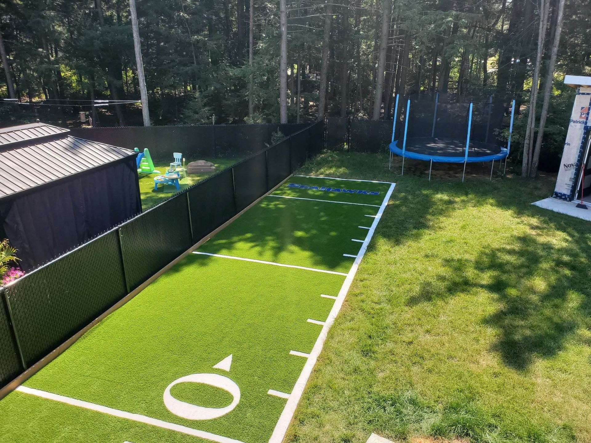 Arrière-cour avec un petit terrain de football marqué sur du gazon vert, un trampoline et une clôture noire, des arbres en arrière-plan.