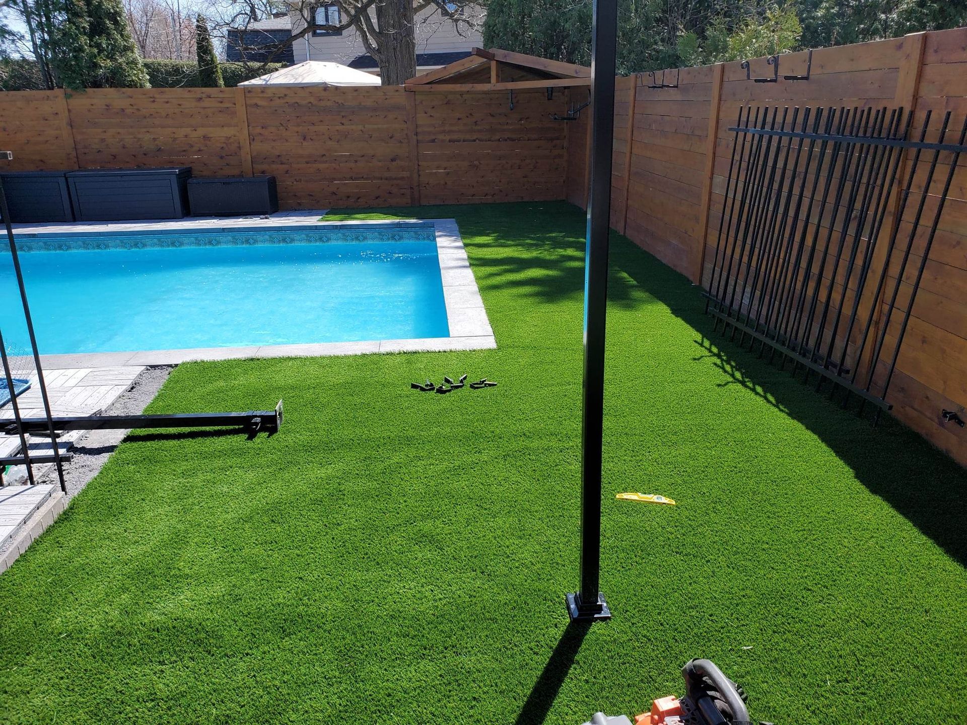 Un jardin avec une piscine rectangulaire, du gazon synthétique et une clôture en bois. Au premier plan, un poteau en métal noir et quelques outils.
