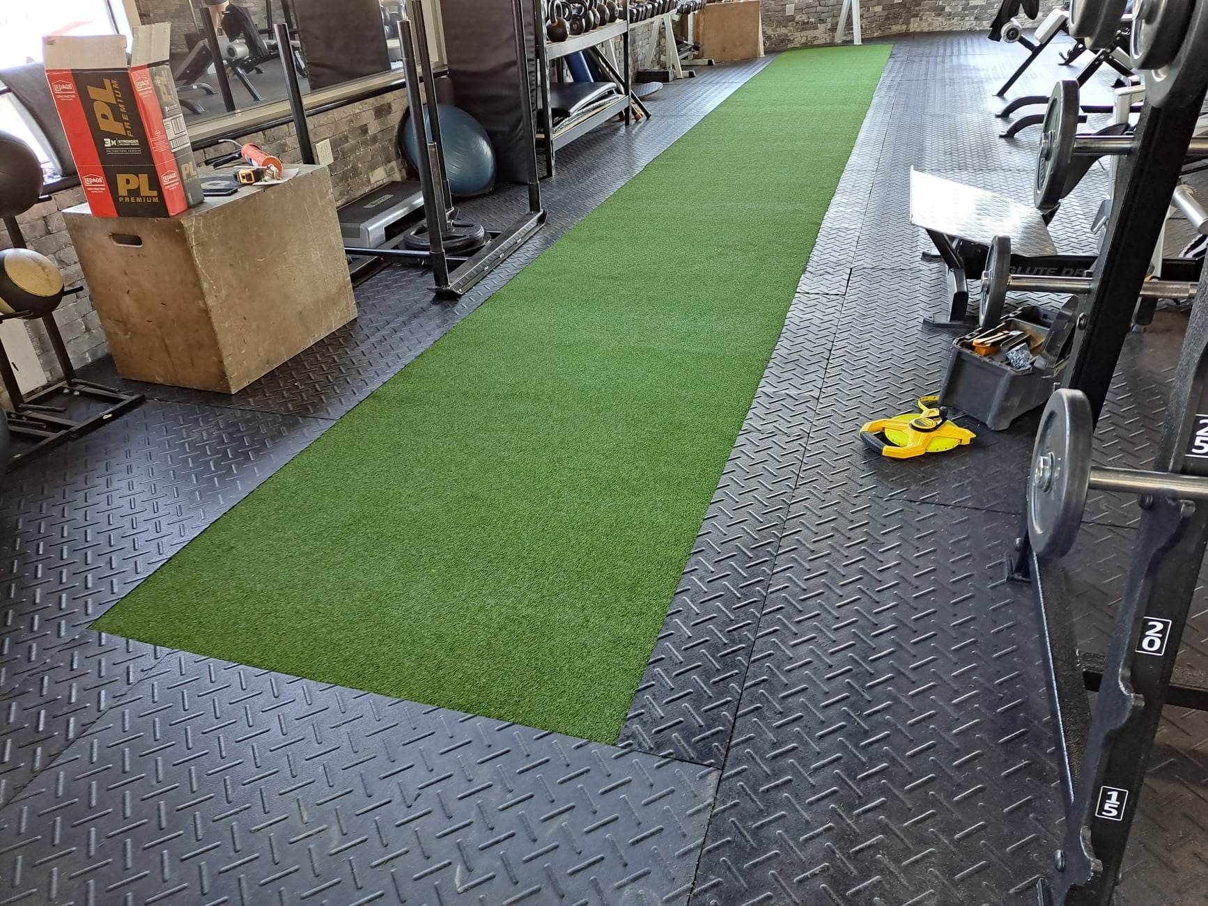 Intérieur de salle de sport avec une bande de gazon vert entourée d'un revêtement en caoutchouc noir, équipement visible de chaque côté.
