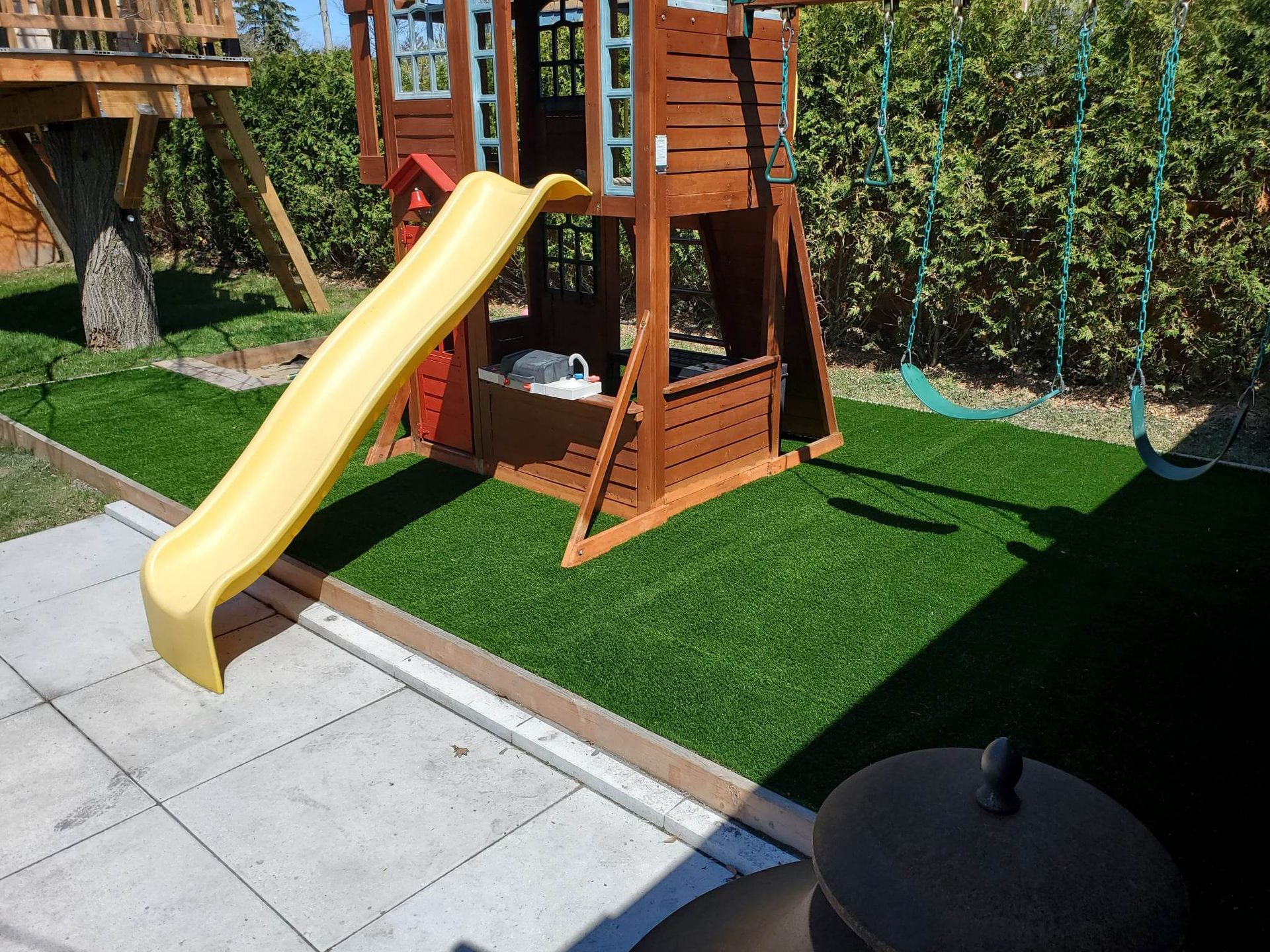 Un ensemble de jeux en bois avec un toboggan jaune et des balançoires est posé sur du gazon vert artificiel dans une cour arrière.