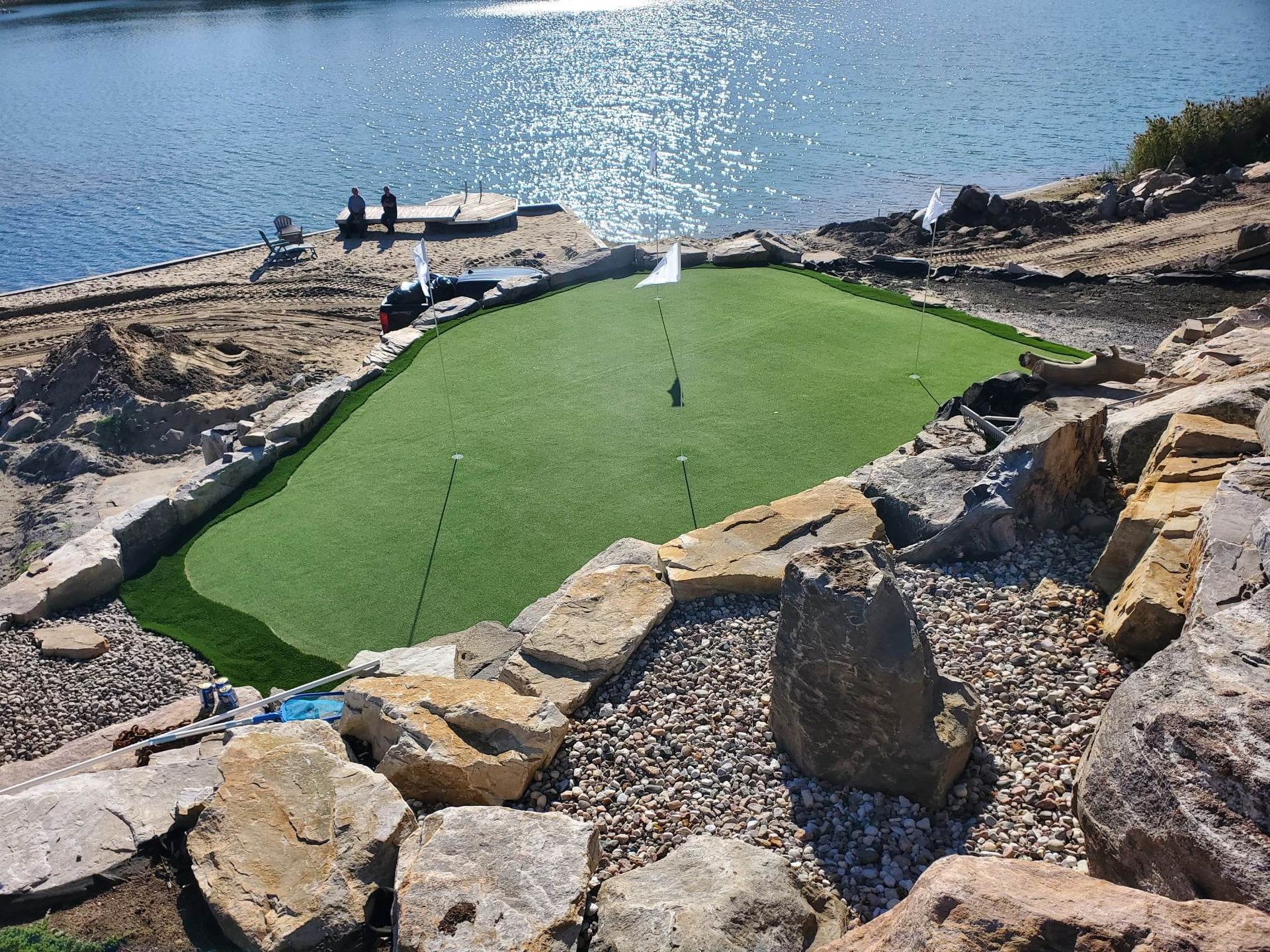 Un green de golf avec un drapeau se trouve au bord d'un lac, encadré de rochers. Deux personnes se tiennent sur un quai en arrière-plan.