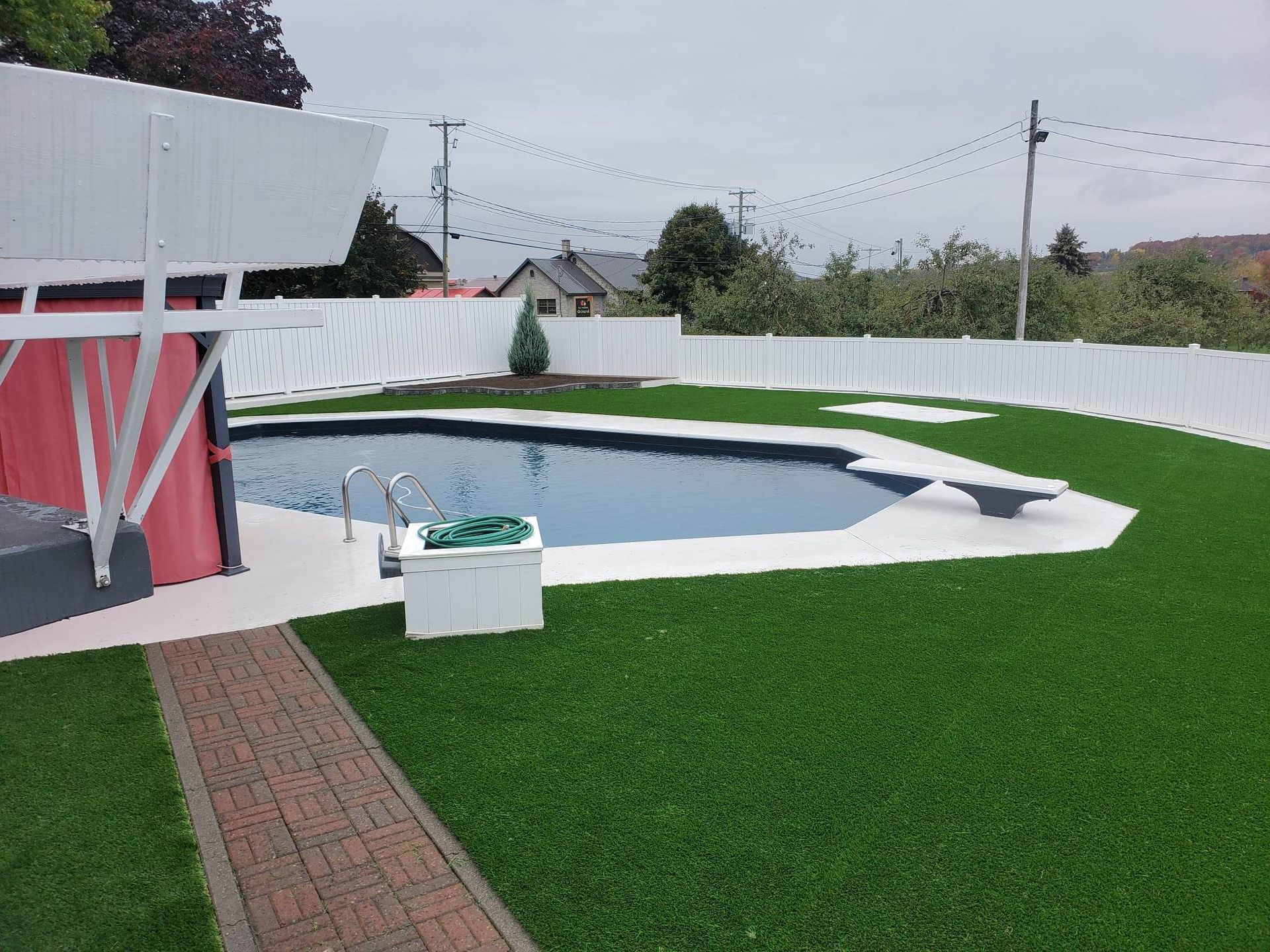 Une piscine avec plongeoir, entourée de gazon vert et d'une clôture blanche. Un bâtiment rouge se trouve sur la gauche, et un chemin en briques mène à la piscine.