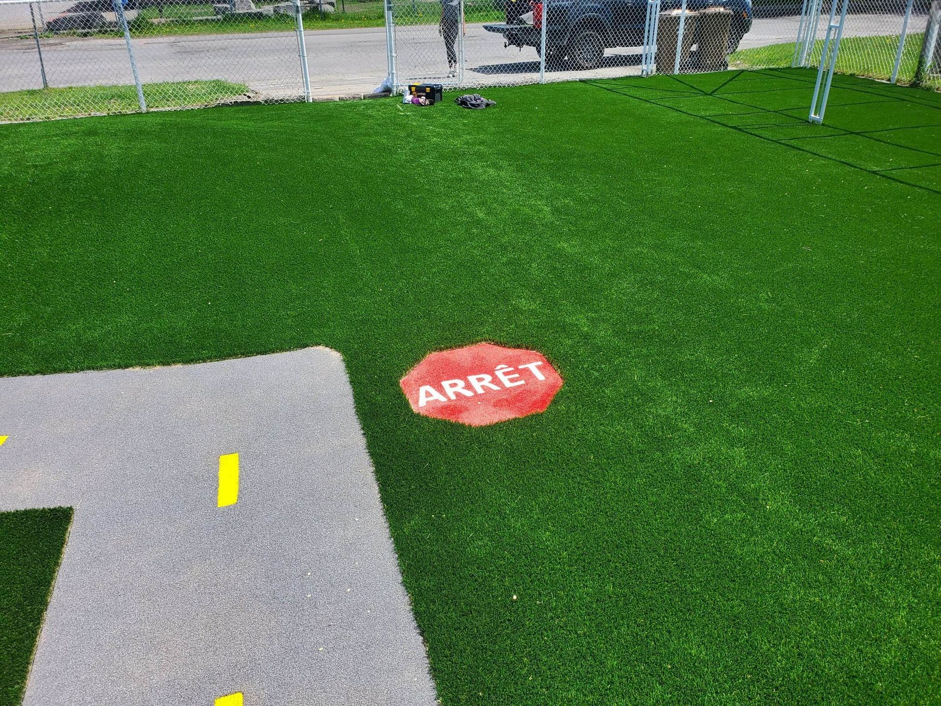 Aire de jeux en gazon synthétique, comportant une zone pavée grise délimitée par des lignes jaunes et un panneau d'arrêt rouge en français, « ARRÊT ».