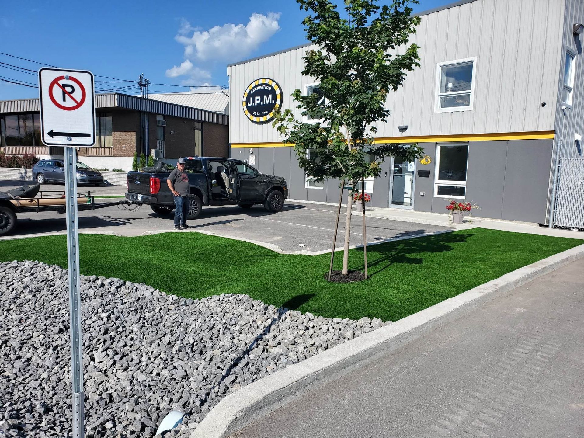 Un homme à côté d'un camion garé près d'un bâtiment avec le logo « JMV », de petites parcelles de gazon artificiel et un panneau d'interdiction de stationnement.