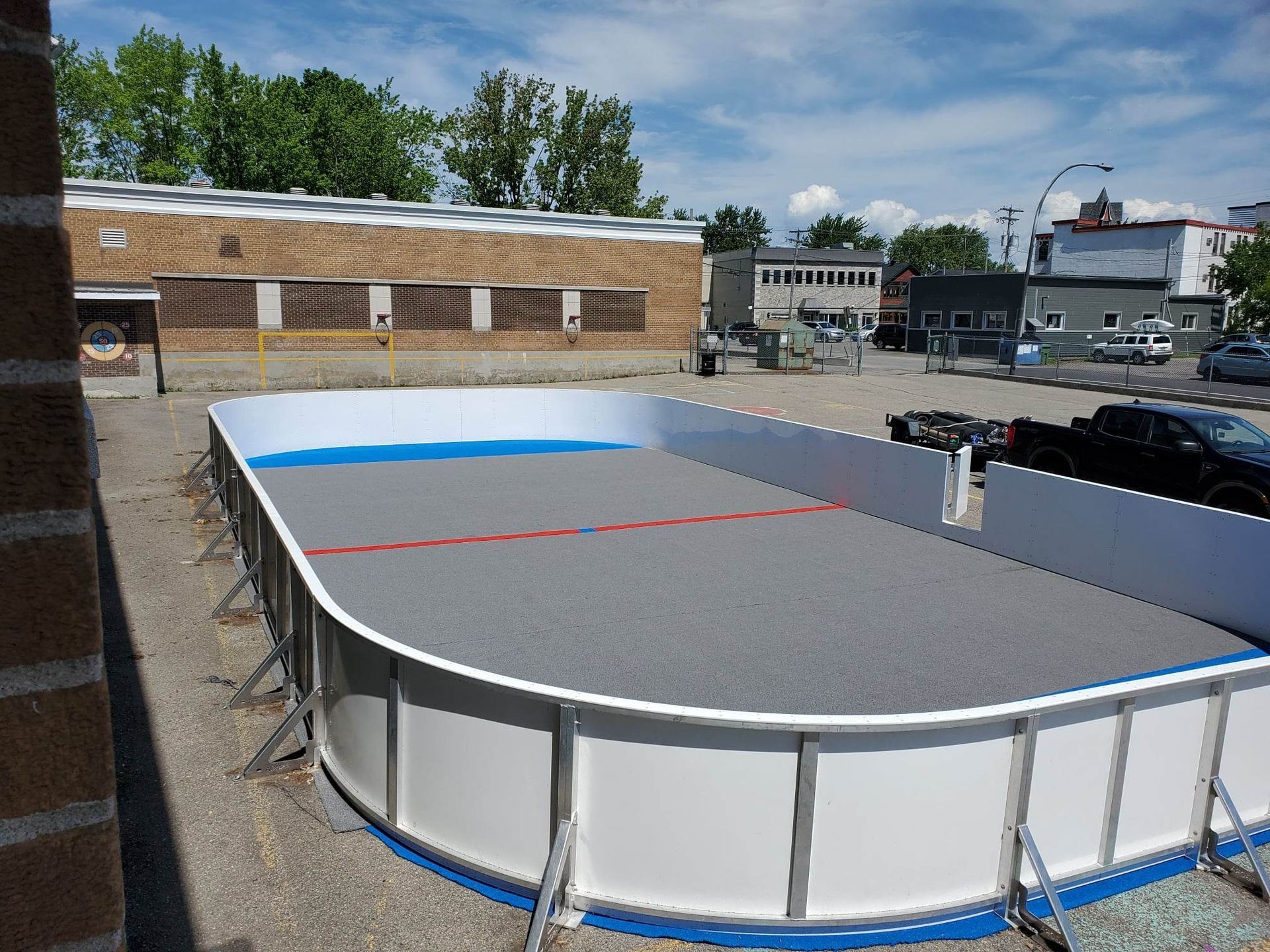 Patinoire de hockey extérieure aux murs bleus, gris et blancs. Un bâtiment en briques et des voitures garées sont visibles en arrière-plan.